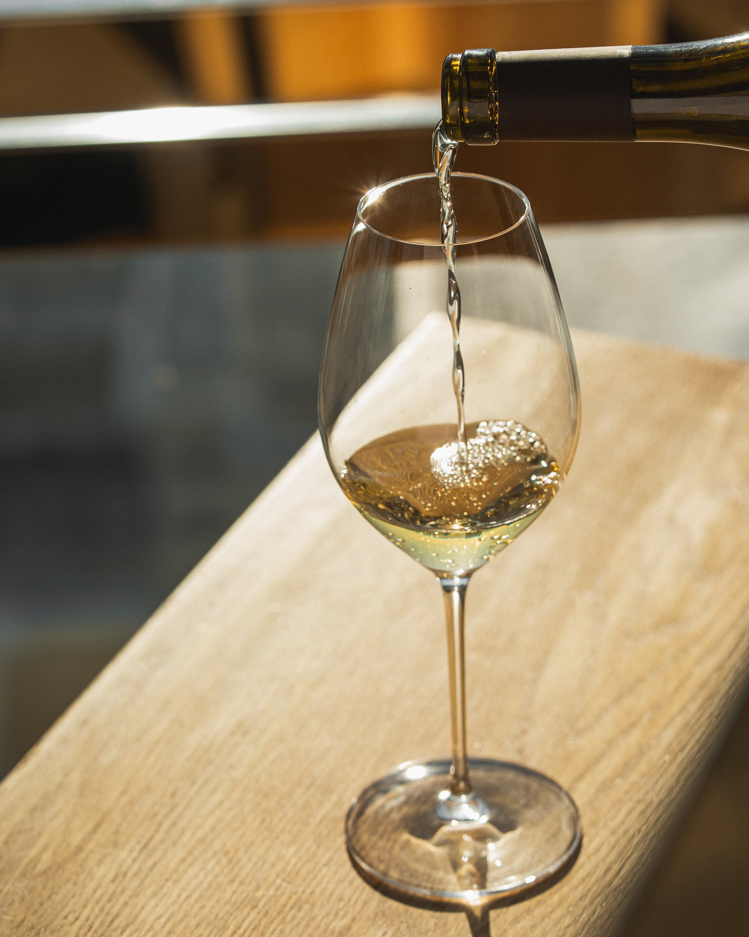 A glass of white wine being poured from a bottle. The glass is on a wooden surface and the wine is mid-pour, creating bubbles and a splash inside the glass.