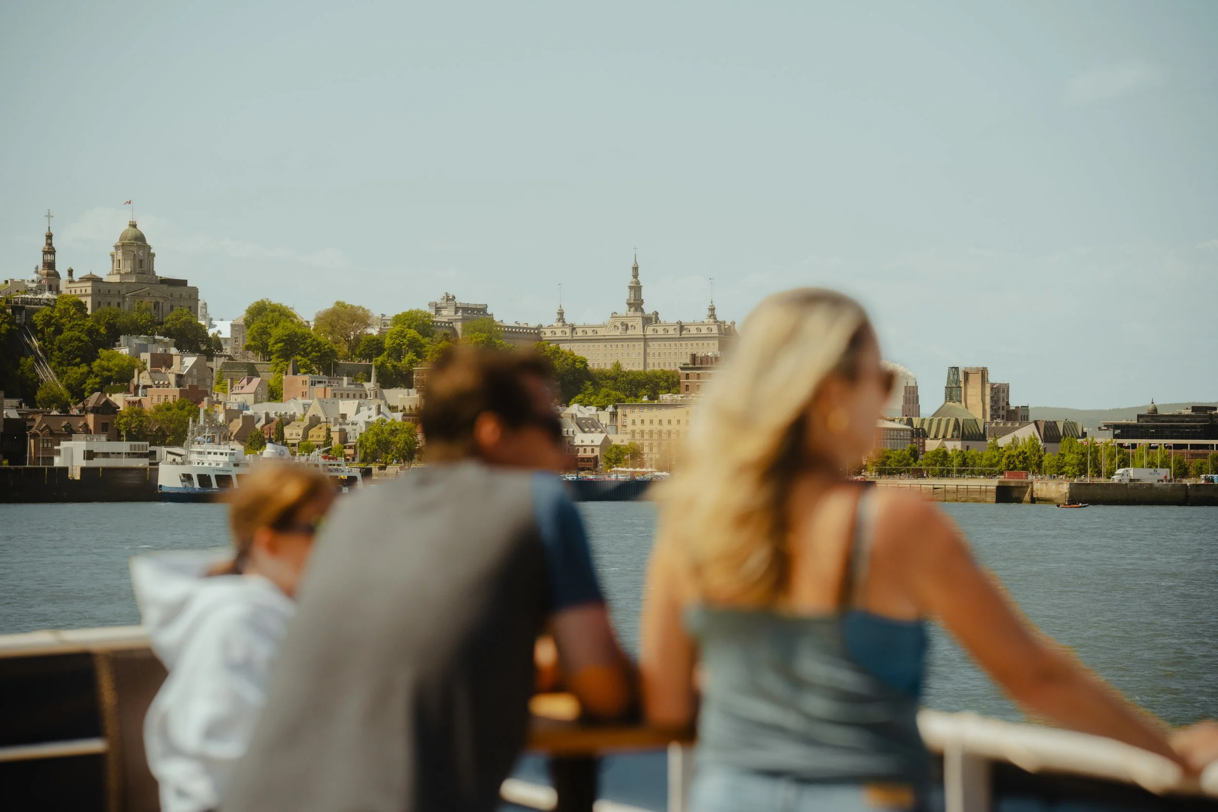 People sitting on a boat or ferry enjoying a river view of a city skyline, with prominent historic buildings and lush green trees on the hills in the background.