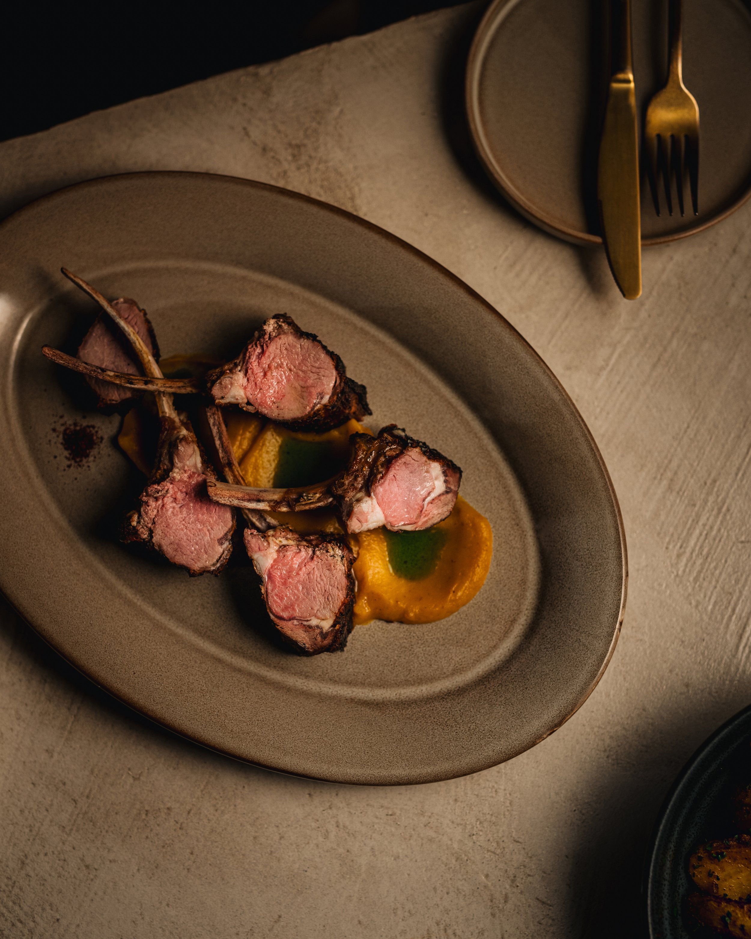 A plate of sliced, cooked lamb chops garnished with herbs and served with a creamy yellow sauce and green sauce underneath.