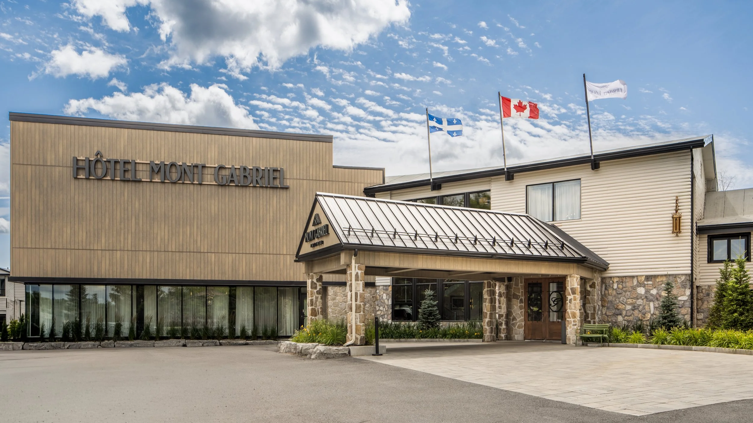 Exterior of Hôtel Mont Gabriel with flags of Quebec, Canada, and Mont Gabriel flying on the building's roof, under a partly cloudy sky.