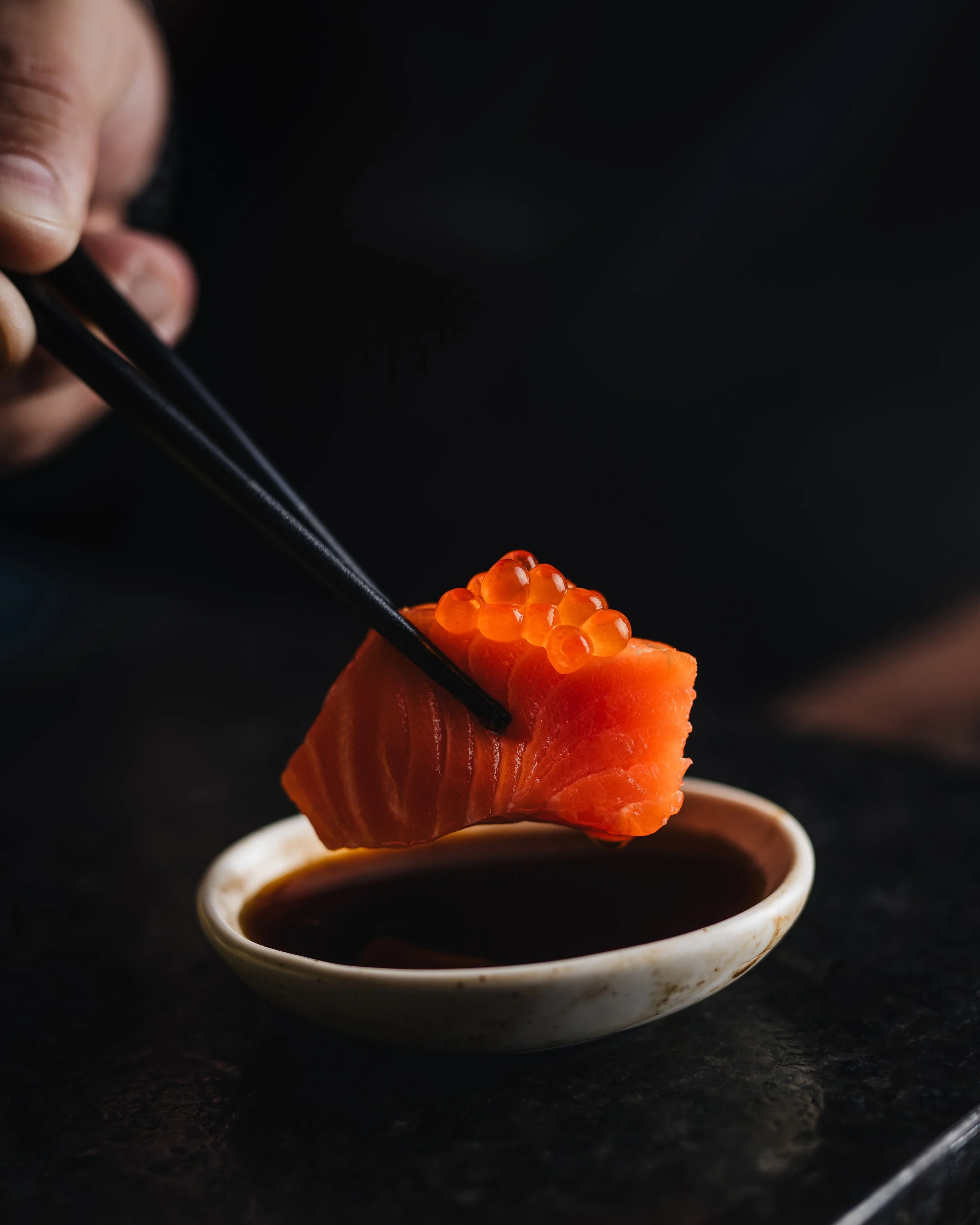 Close-up of a hand holding chopsticks carefully lifting a piece of raw salmon sashimi topped with orange salmon roe over a dish of soy sauce on a dark surface.