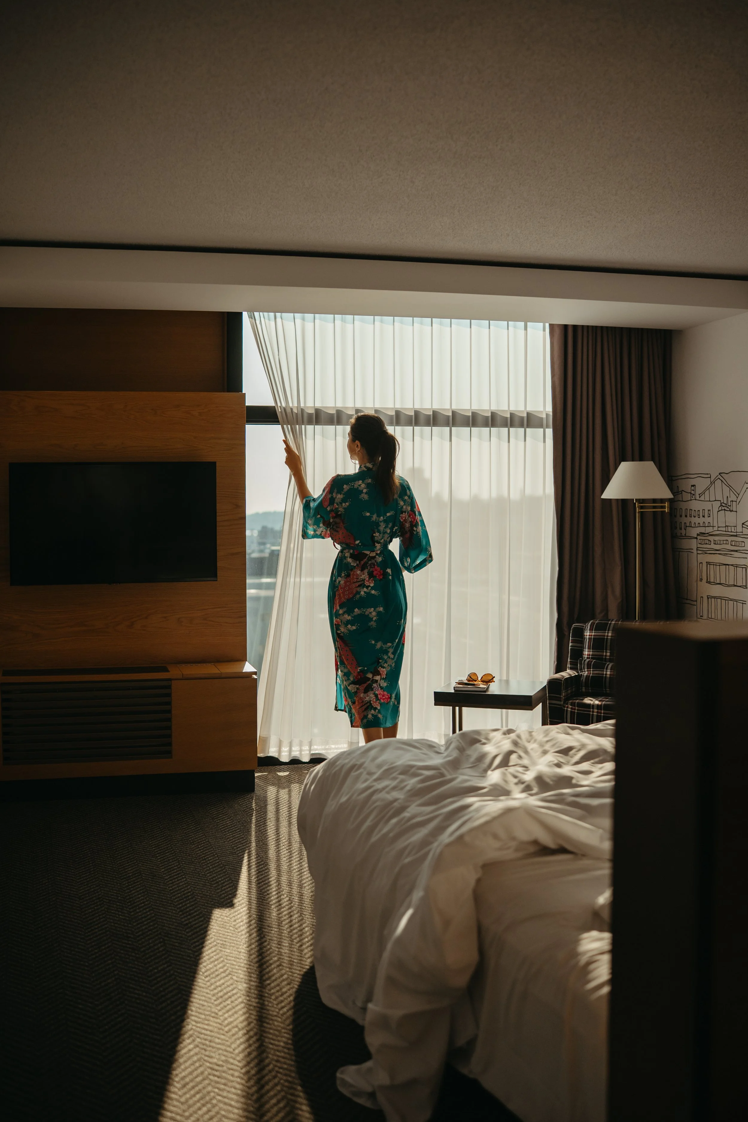 A woman wearing a floral kimono standing by a window, partially open, with curtains pulled to the side, in a hotel room. The bed is unmade in the foreground, with a nightstand and lamp beside the window.
