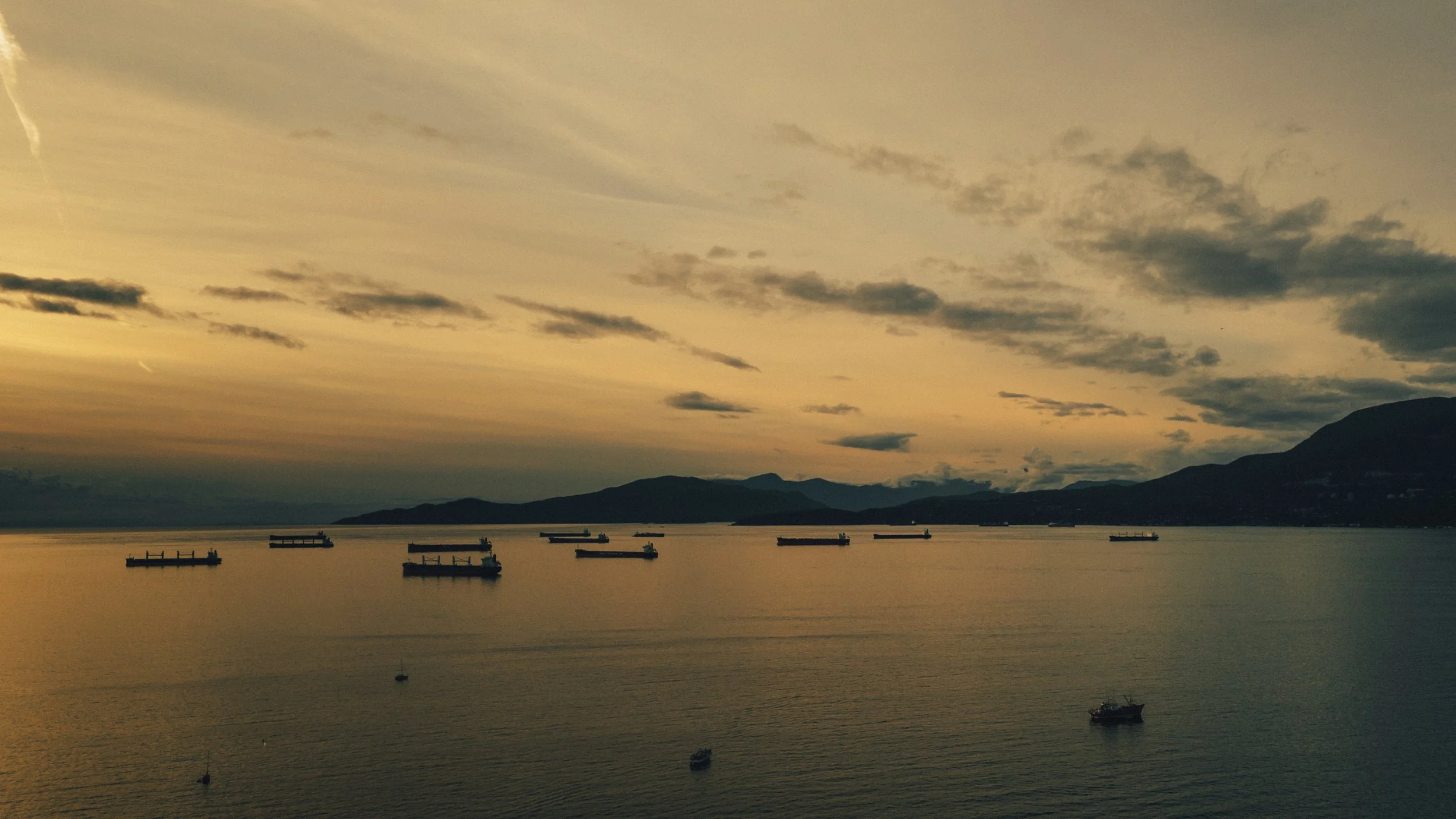 A serene body of water at sunset with multiple cargo ships anchored across the calm water, mountains in the distance under a sky with scattered clouds.