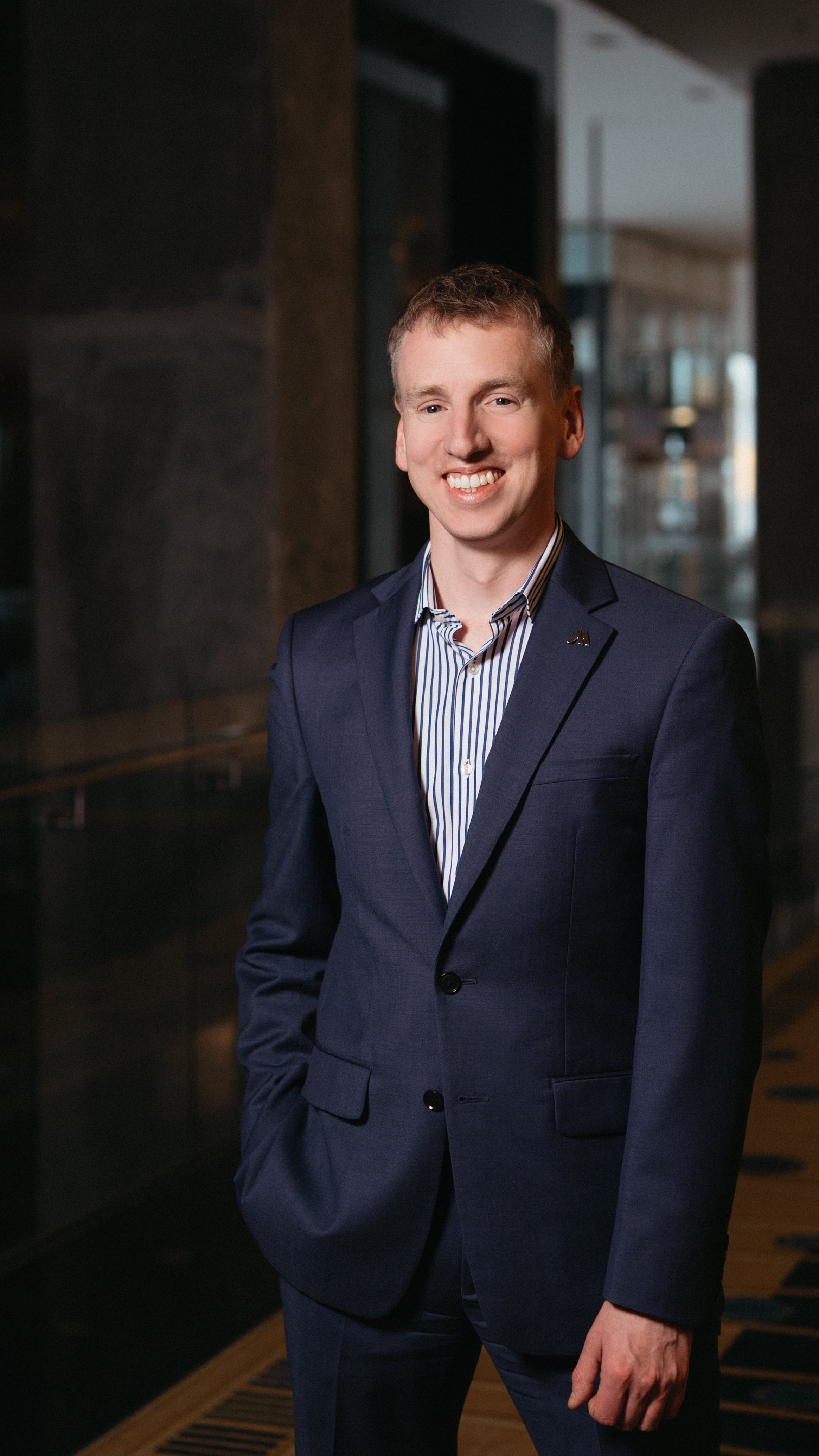 A young man with short light brown hair, wearing a navy blue suit with a striped dress shirt, standing indoors in front of a dark background and windows, smiling at the camera.