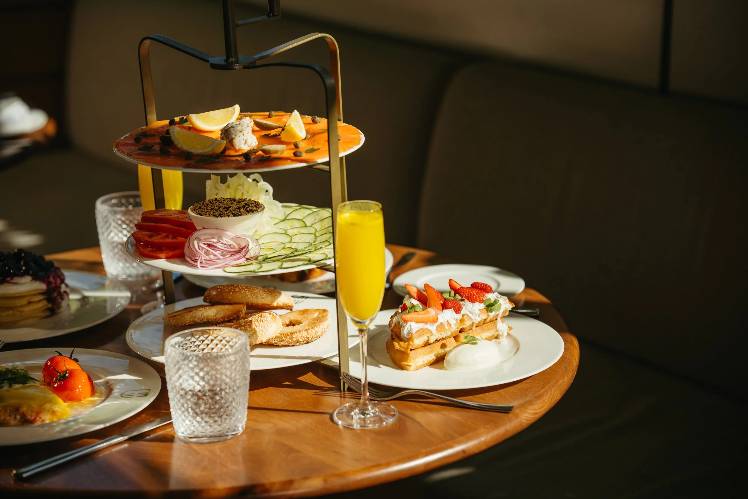 A multi-tiered tray of assorted foods including smoked salmon with lemon wedges, sliced cucumbers, tomatoes, cheese, bread, and desserts like waffles with strawberries and whipped cream, set on a table with glasses of orange juice.