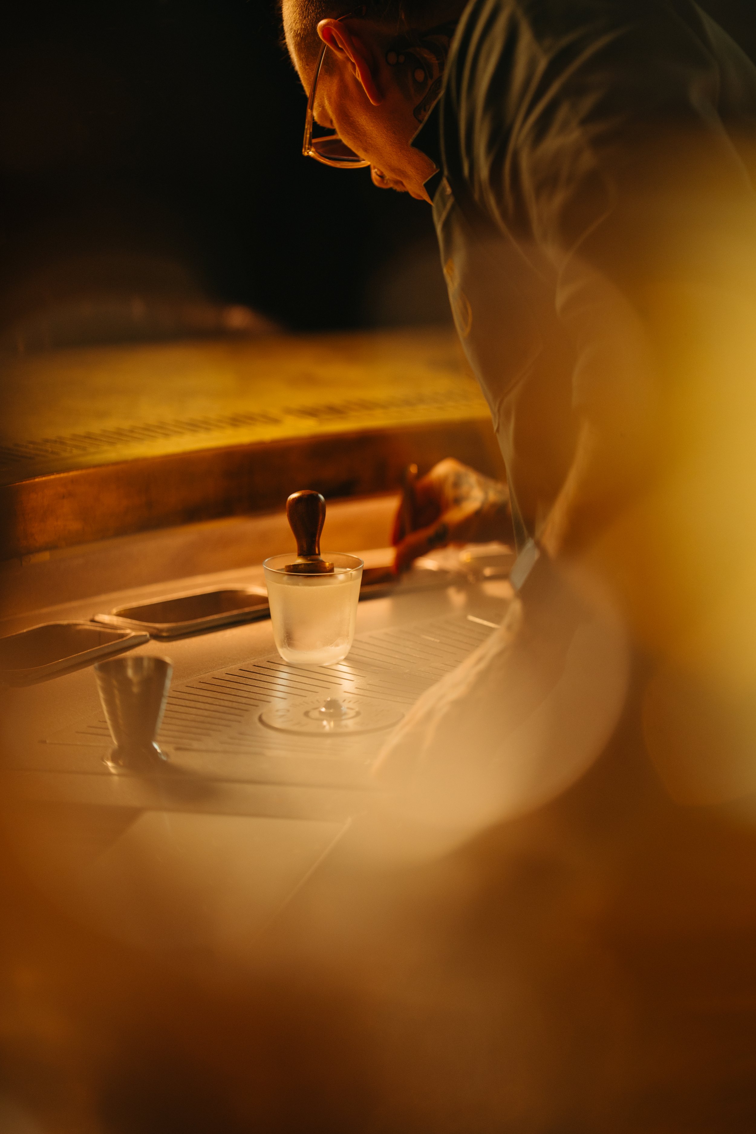 A person with glasses and tattoos on their neck, wearing a striped shirt, is leaning over a table with a candle and some equipment, in a warm, dimly lit environment.