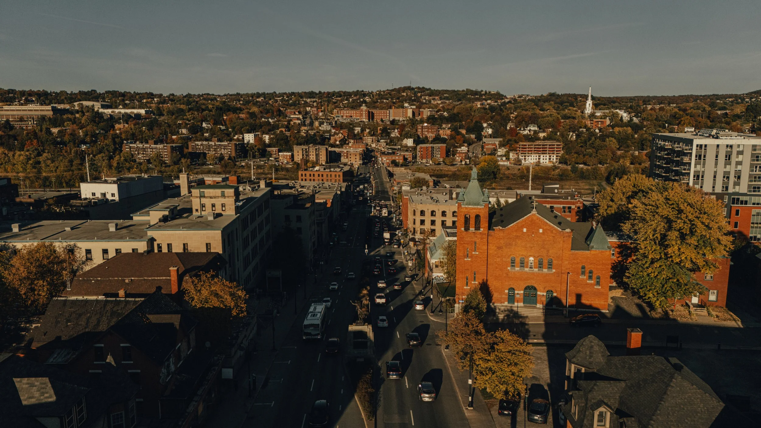 A city view with buildings, streets, trees with fall foliage, and a church with a tall steeple in the foreground.