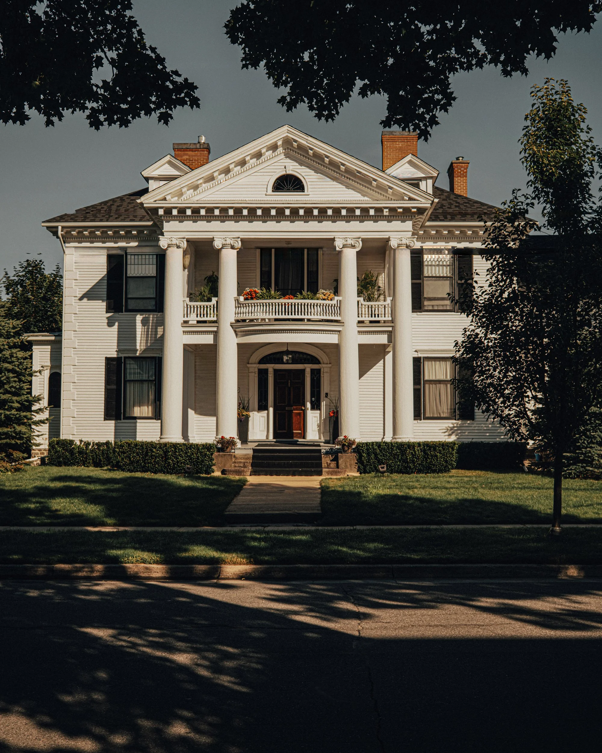 Front view of a large, white, classical-style house with four columns supporting the porch roof. The house has a pitched roof with chimneys and a small arched window on the upper floor. The house is framed by trees and a grassy yard, with shadows cas