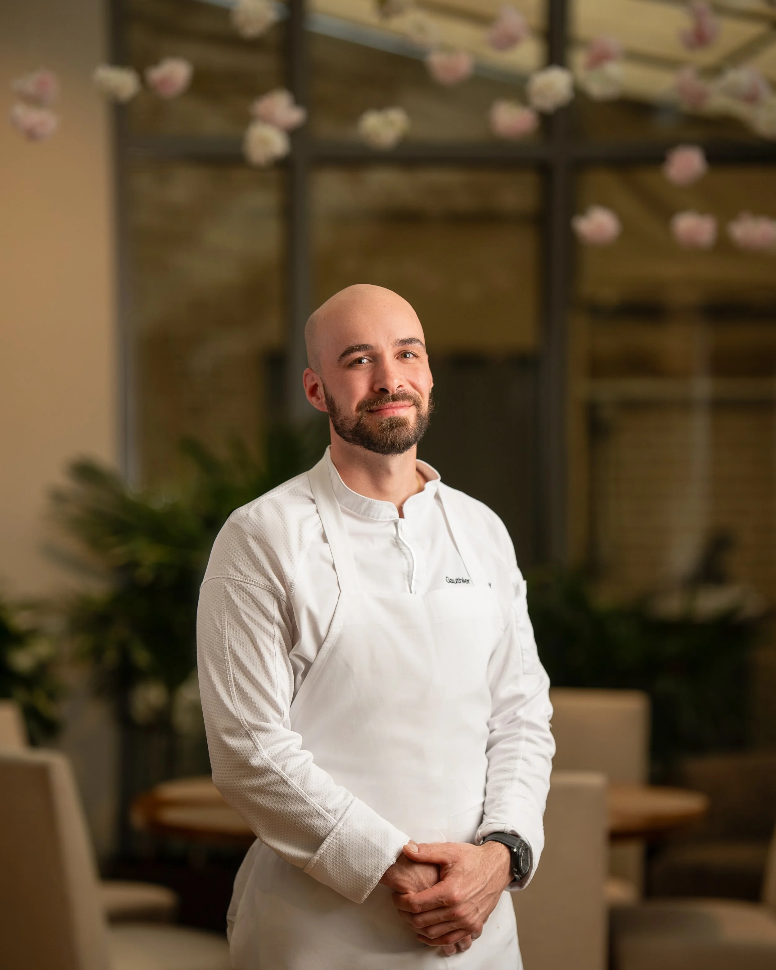 A man wearing a white chef's coat and a watch is standing with his hands clasped in front of him, smiling slightly, in a setting with blurred shelves and pink flowers in the background.
