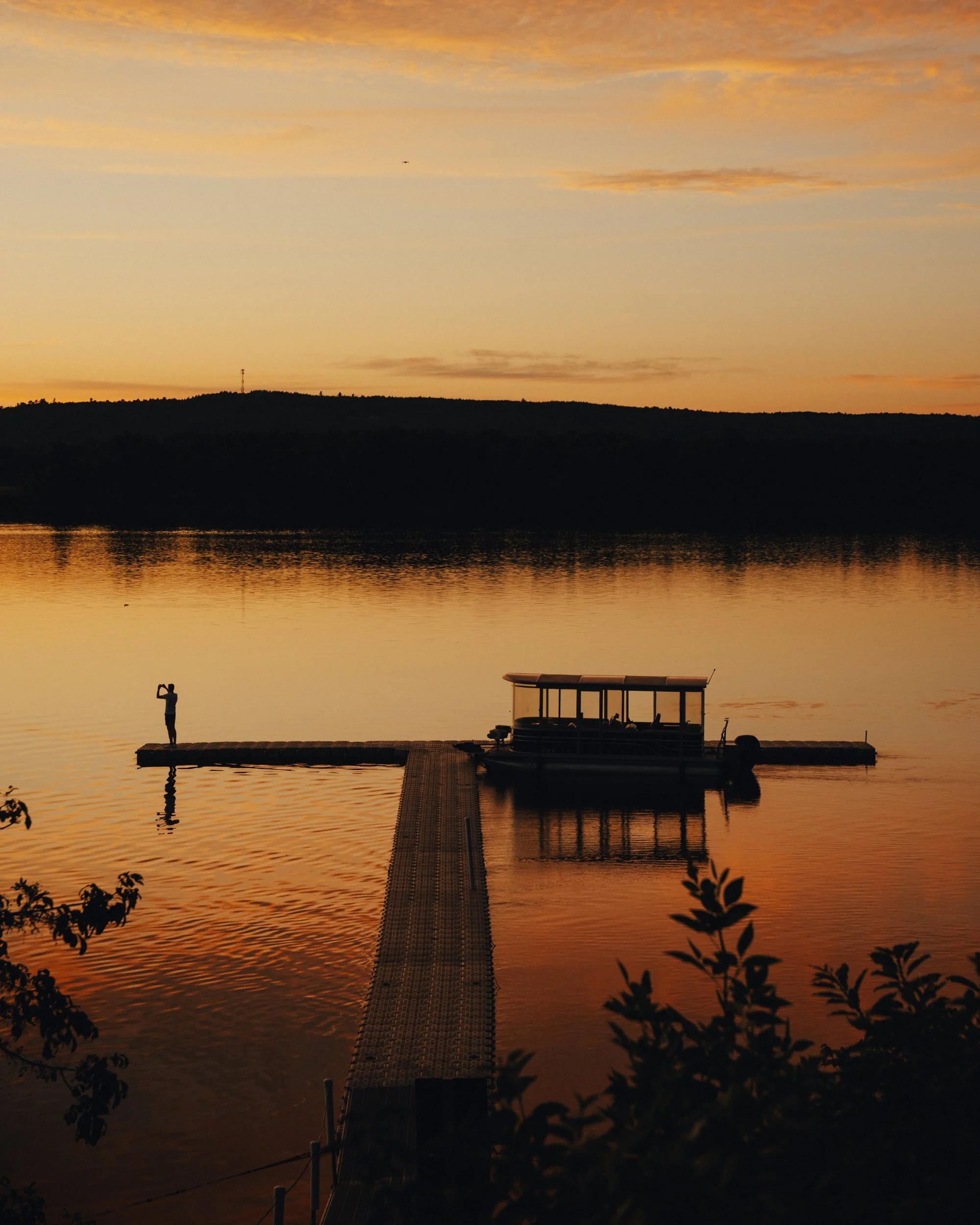 Sunset over a lake with a silhouette of a person standing on a dock and a boat anchored nearby, with trees and hills in the background.