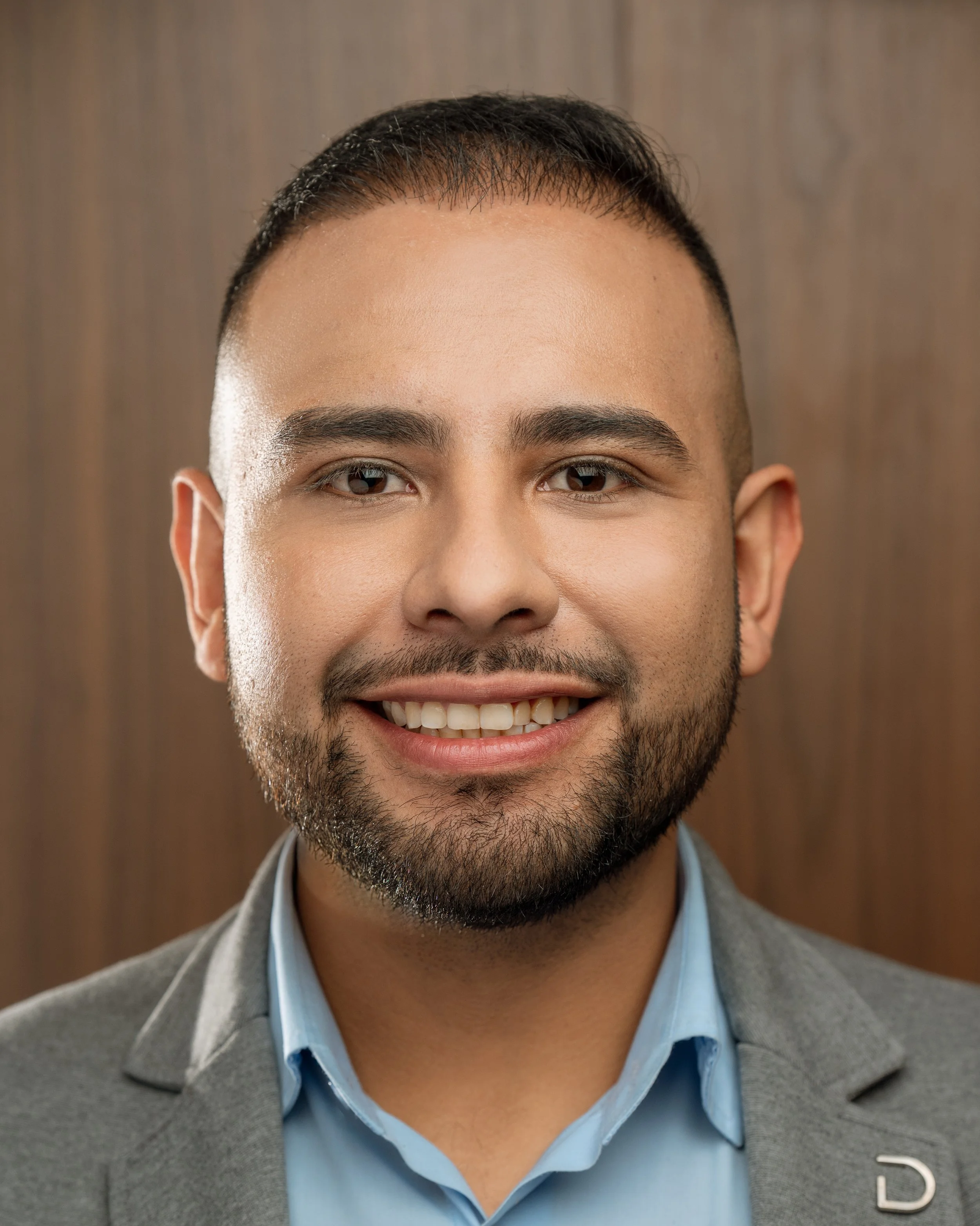 Close-up of a smiling man with dark hair, a beard, wearing a light blue shirt and a gray blazer.