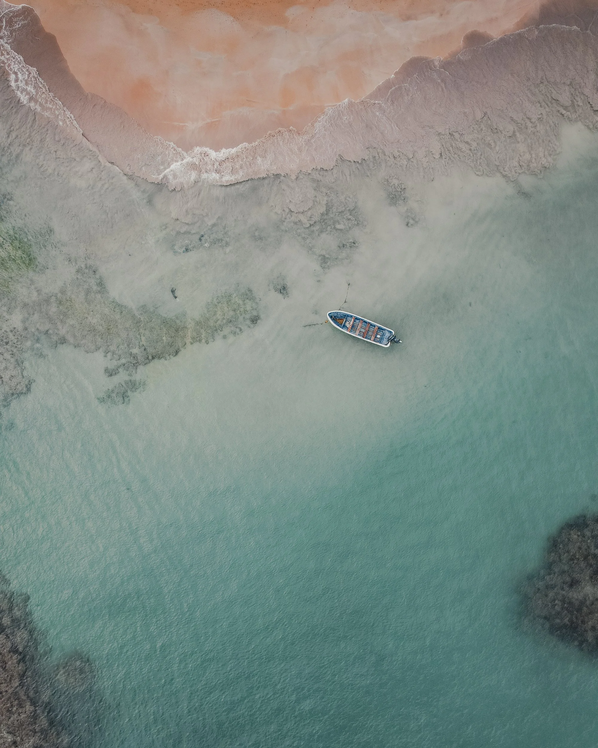 Aerial view of a boat on calm turquoise sea near sandy shoreline with rocky areas and gentle waves.