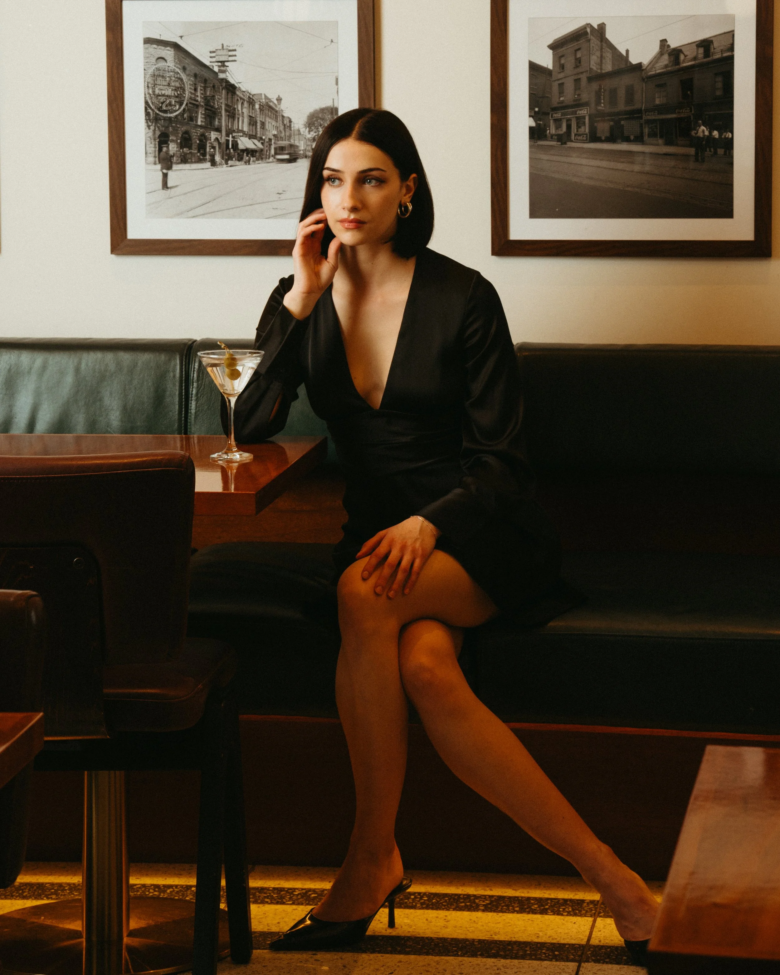 A woman with dark hair and blue eyes sitting at a restaurant table. She is wearing a black dress and large gold hoop earrings, and has a martini with green olives in front of her. She is touching her face with her left hand and looking to the side. B