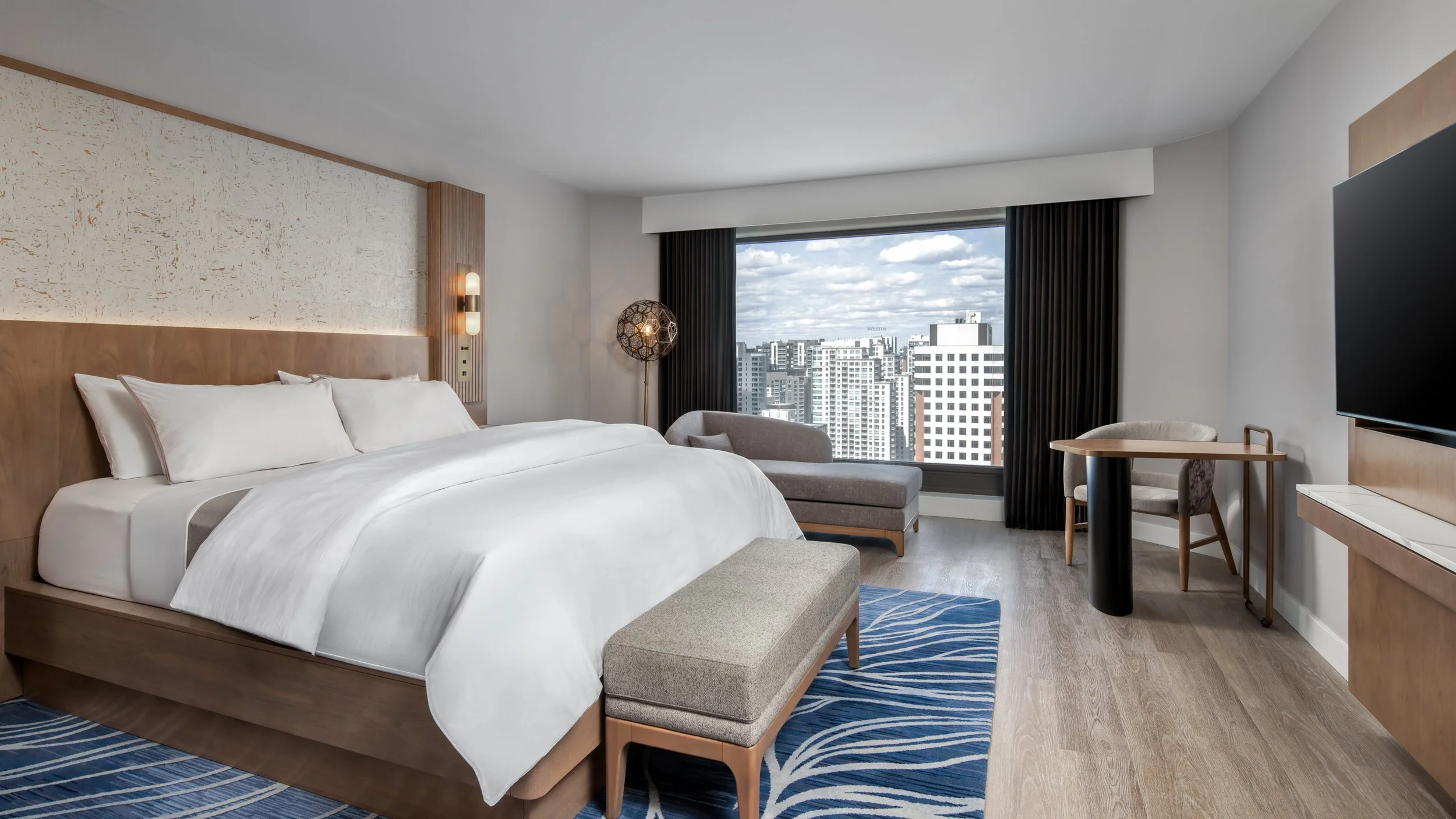 Modern hotel room with a king-sized bed, large window showing city buildings, a sitting area with a chair and ottoman, and a wall-mounted TV.