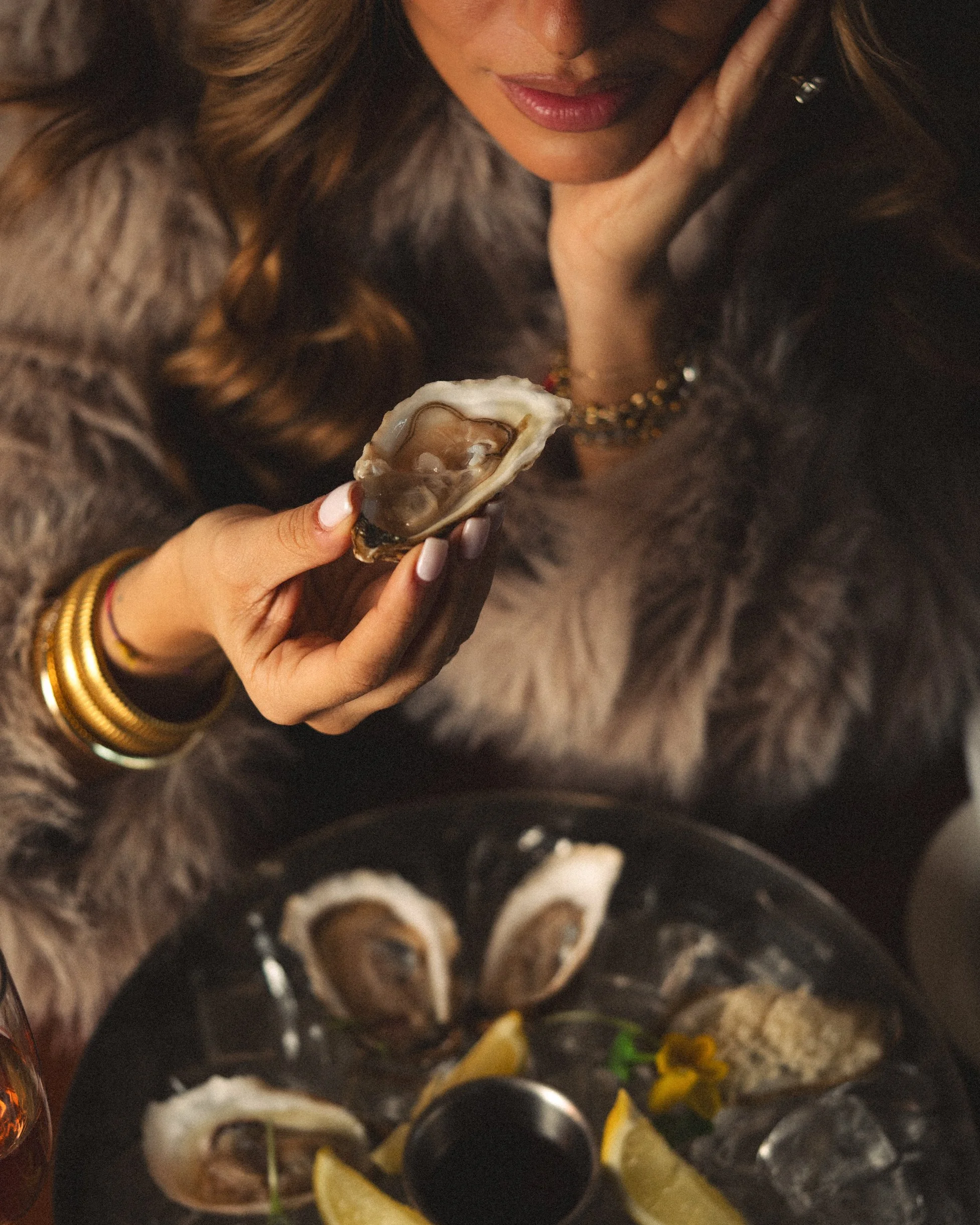 A woman with long brown hair wearing jewelry, holding an oyster shell with a pearl, surrounded by other oysters, lemon wedges, and garnishes on a table.