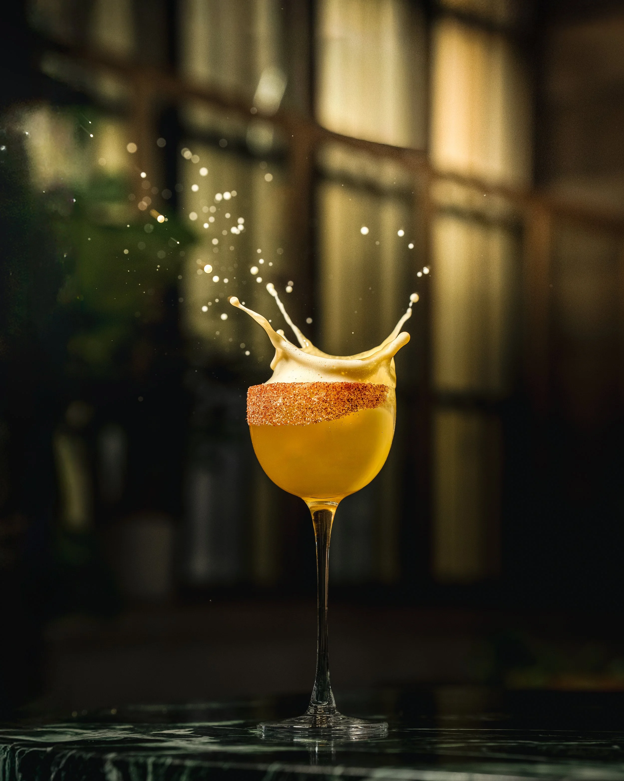 A splash of yellow liquid in a cocktail glass with a sugar-rimmed edge, against a dark, blurred background.