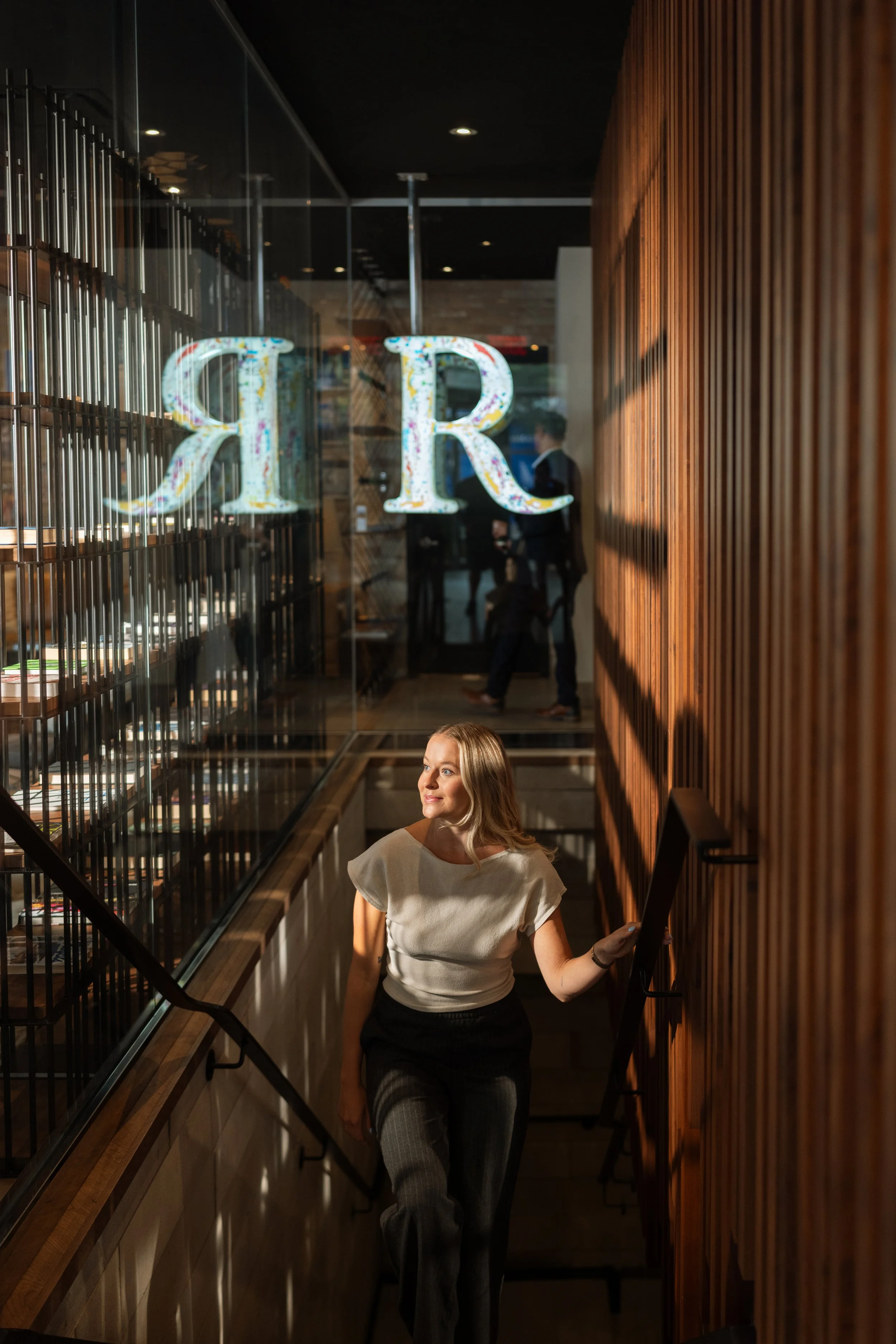 A woman with blonde hair, wearing a beige top and gray plaid pants, walking down a staircase in a modern interior with wooden wall panels and a glass display case.