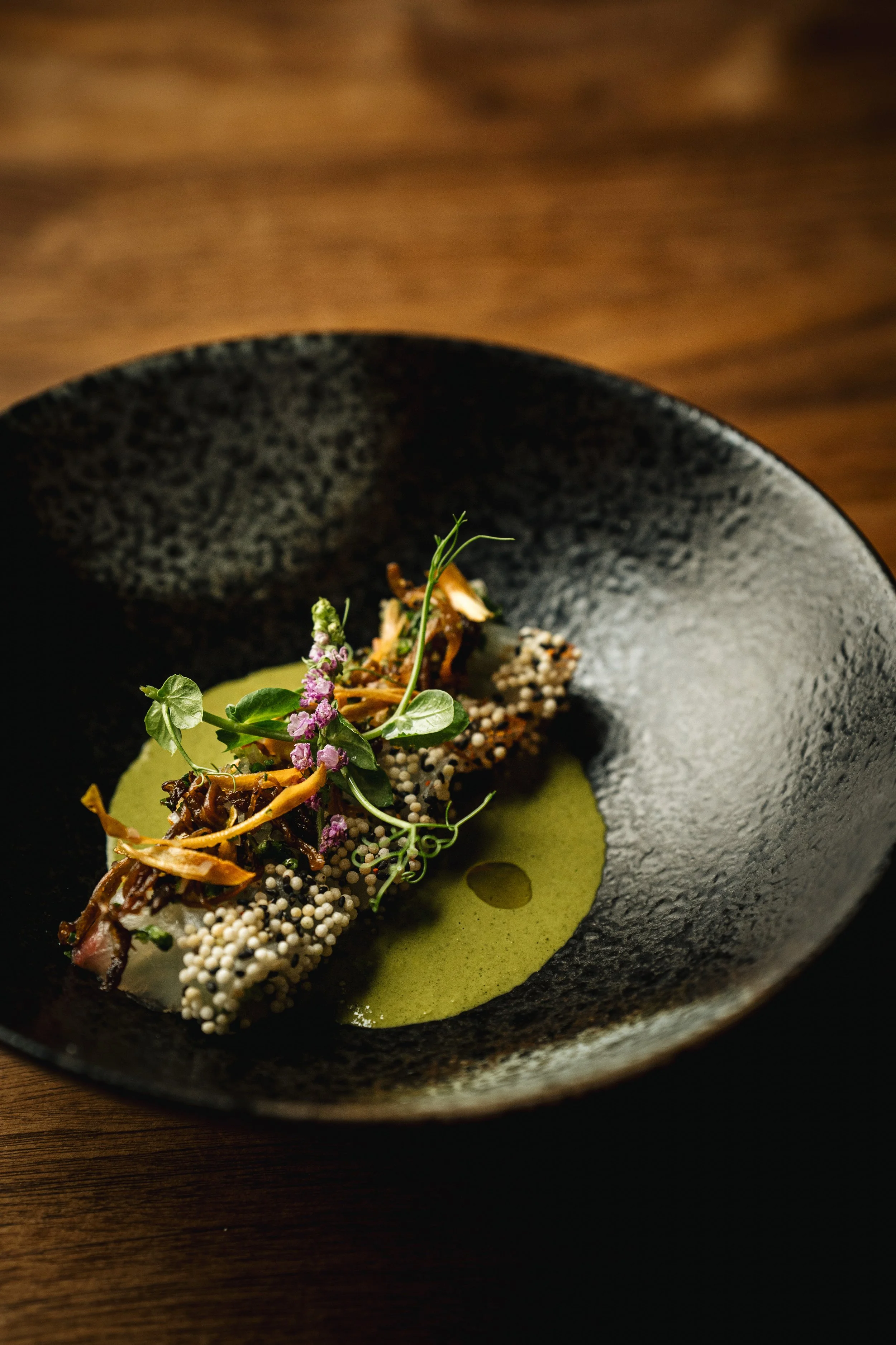 A plated gourmet dish with a green sauce, garnished with herbs, microgreens, purple flowers, and crispy toppings, served in a black textured bowl on a wooden table.