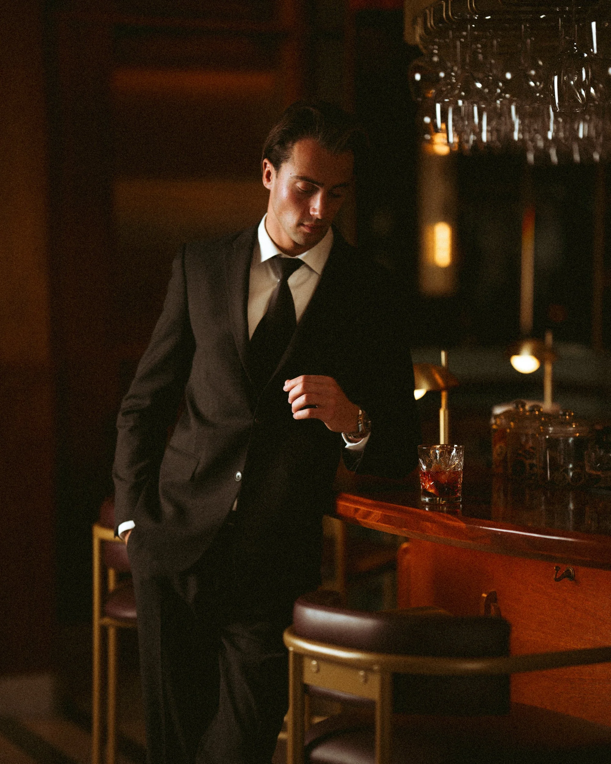 A man in a black suit, white shirt, and black tie standing in a dimly lit bar or restaurant, with a glass of dark beverage on the table in front of him, and a rack of hanging glasses in the background.