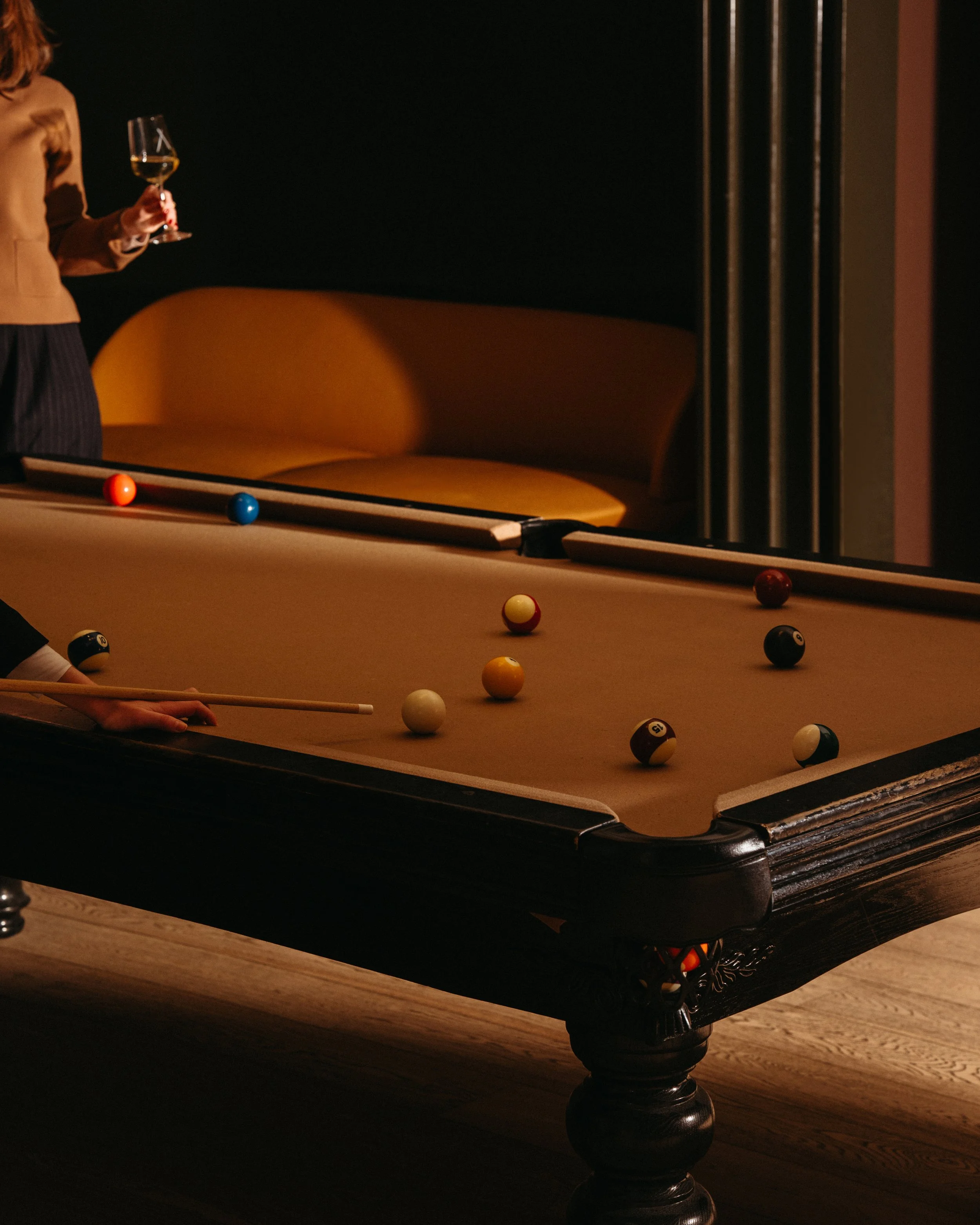 A billiards table with several balls scattered on it, a person holding a cue stick, and a woman holding a glass of white wine. There is a black wall and a yellow couch in the background.