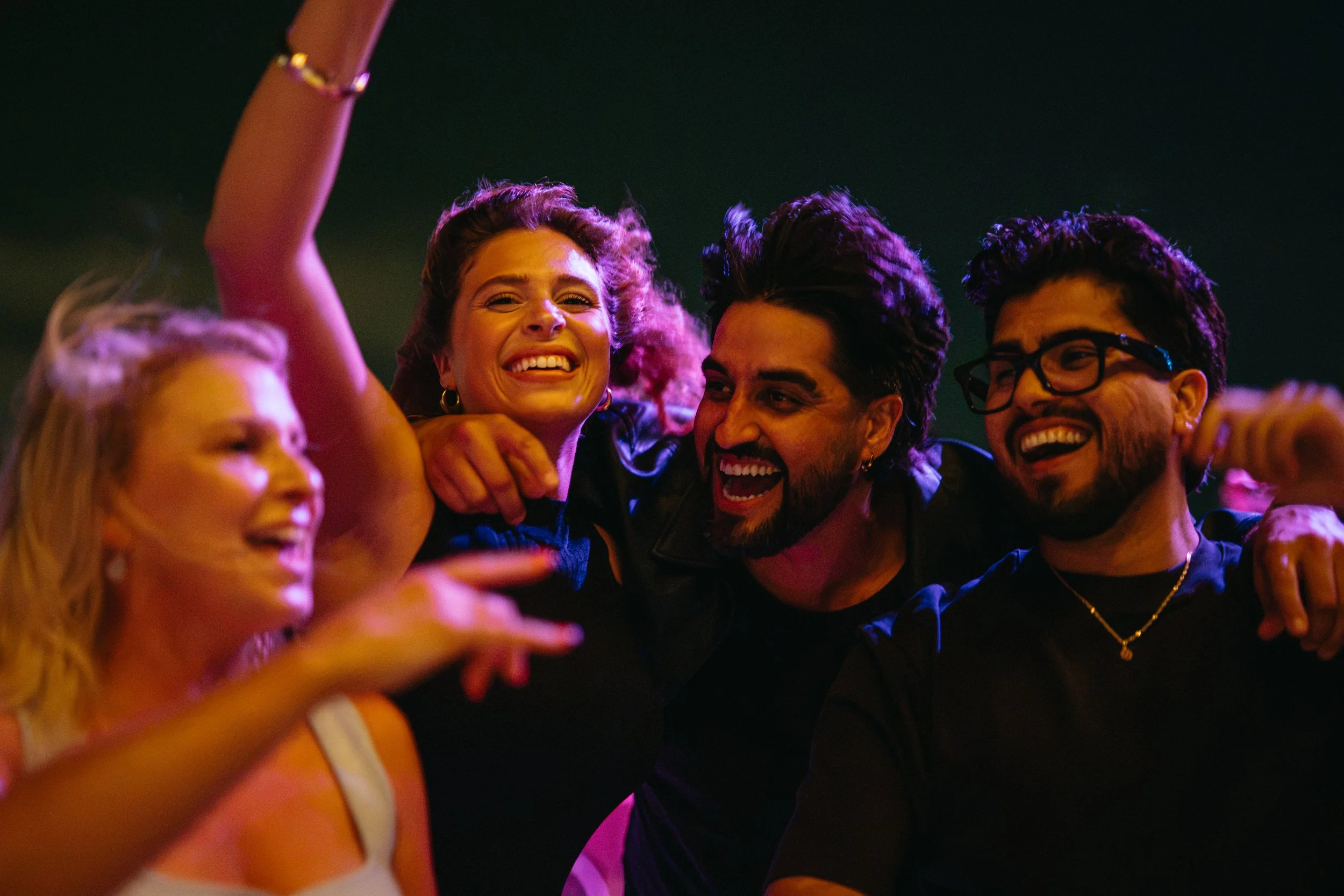 Group of young adults enjoying a night out, smiling, hugging, and taking a selfie together in a dimly lit setting.