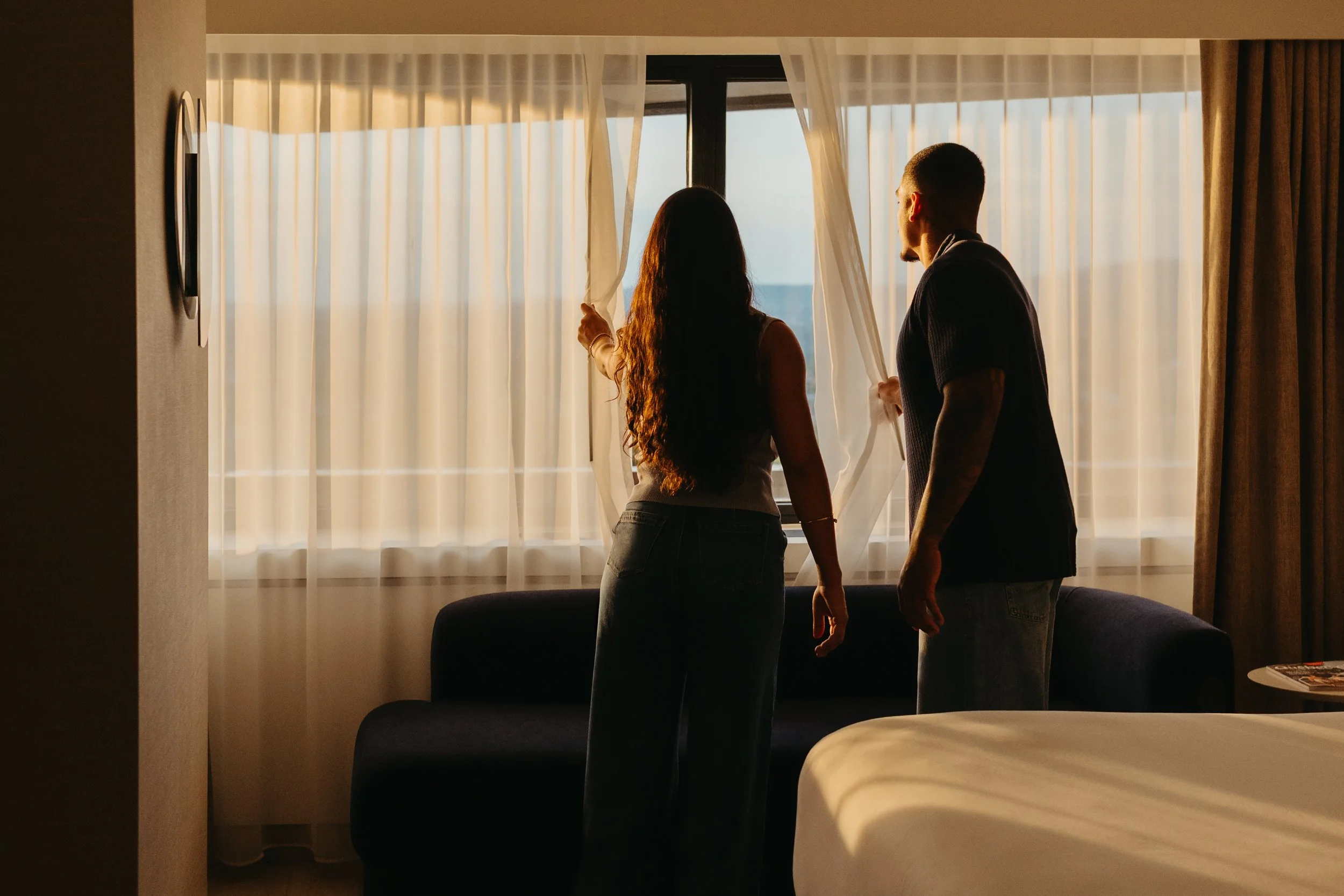 A man and woman are standing inside a hotel room, looking out the window at sunset or sunrise through white curtains. The woman has long, curly hair and is holding back the curtain, while the man is wearing glasses and has short hair.