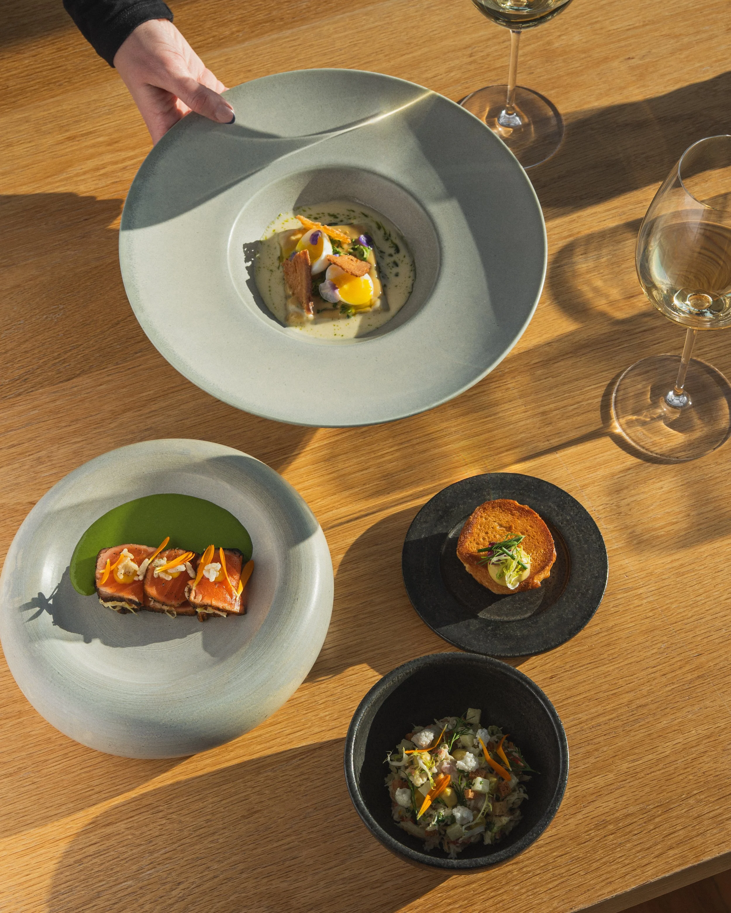 A top-down view of a wooden table with several plated dishes of gourmet food and two glasses of white wine. One person is lifting a greenish-gray bowl containing an elegant dish garnished with herbs and edible flowers.