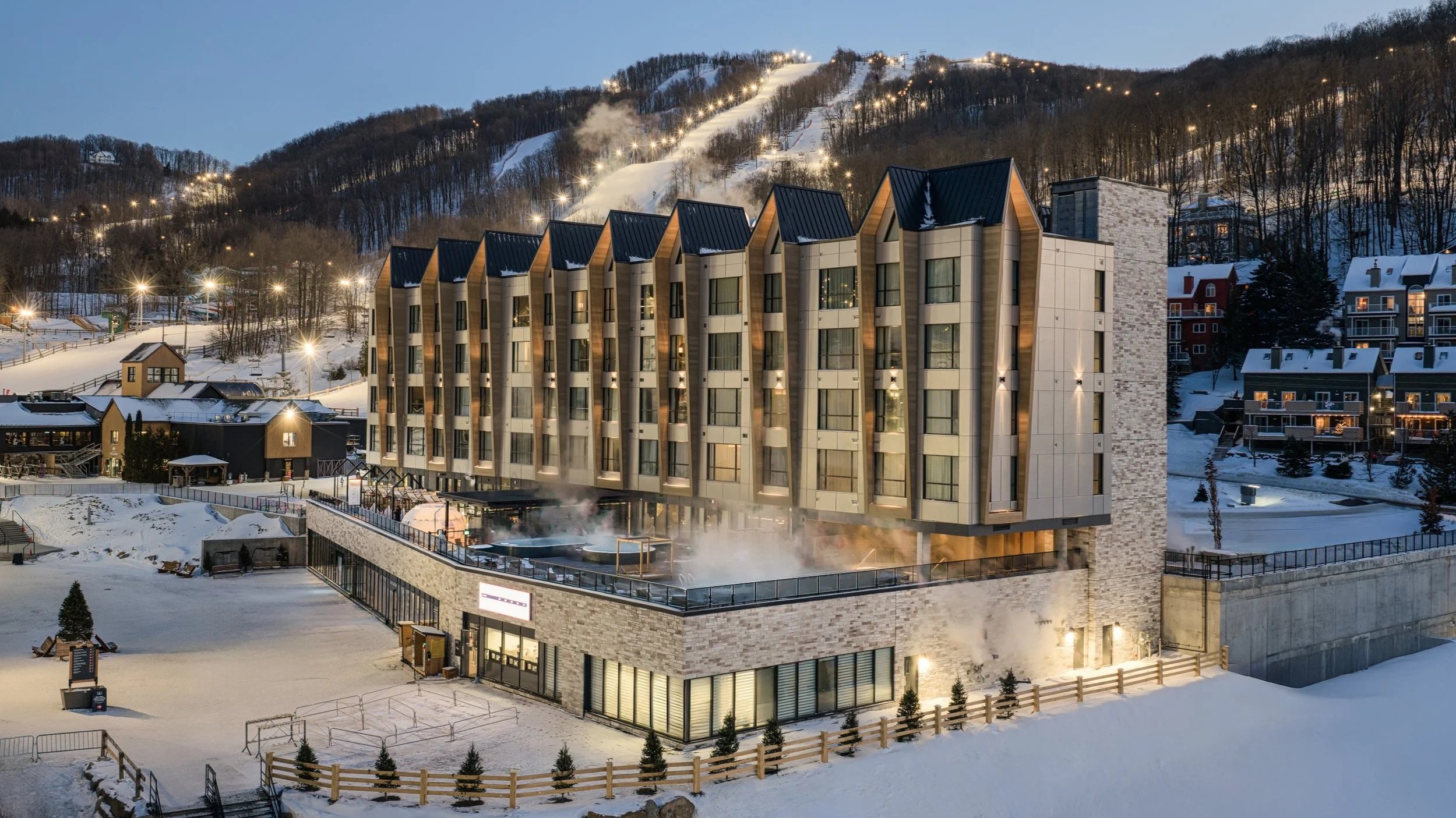 A modern multi-story hotel in a snowy mountain area during dusk, with lights on and steam rising from a hot tub on the terrace.