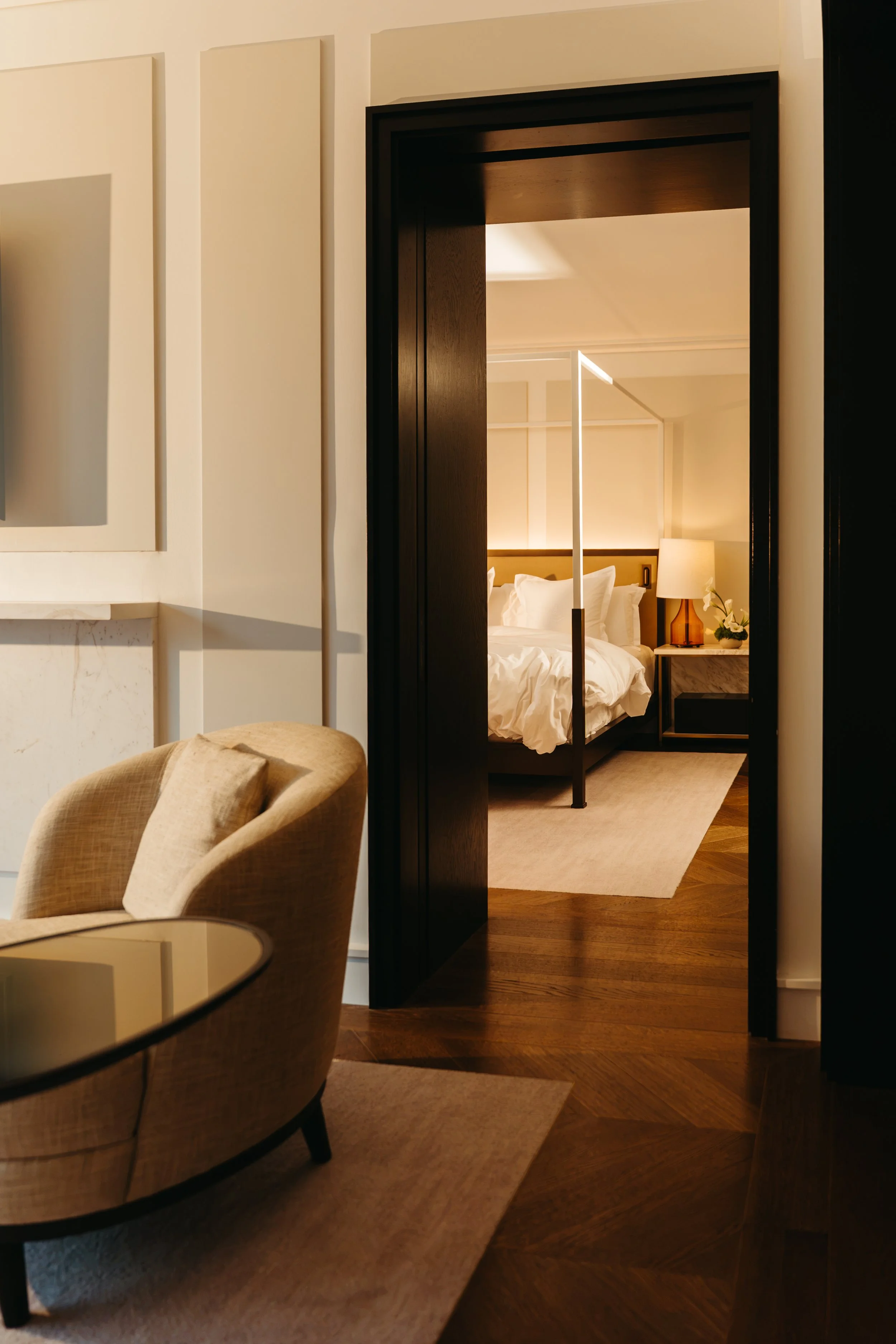 A hotel room with a bed, lamp, and decorative items seen through a doorway, with a dining area with a beige armchair and a glass table in the foreground.