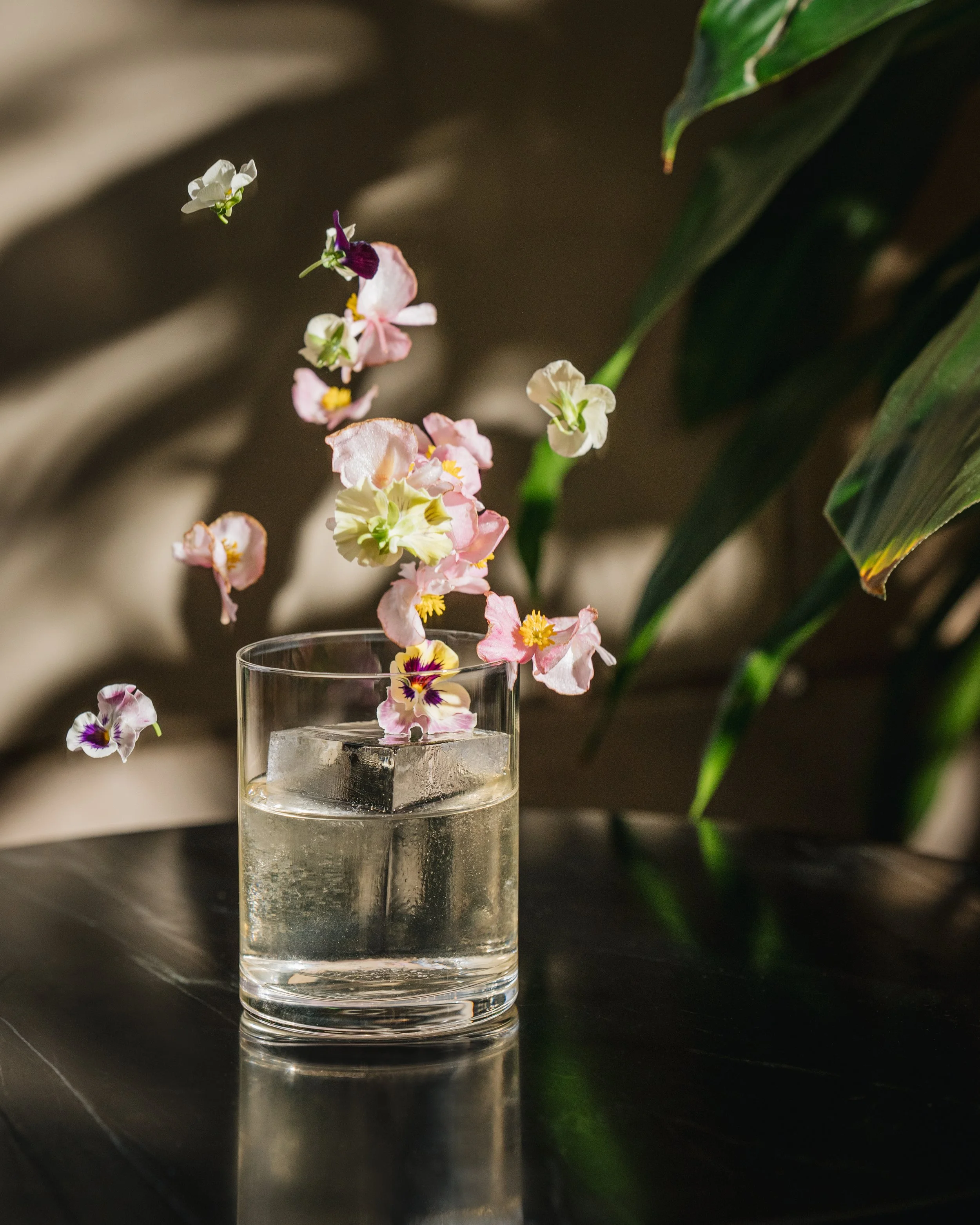A clear glass of water with ice cubes on a dark surface, with a flower arrangement in the background. Some flowers are falling into the glass, and there are green leaves in the background.