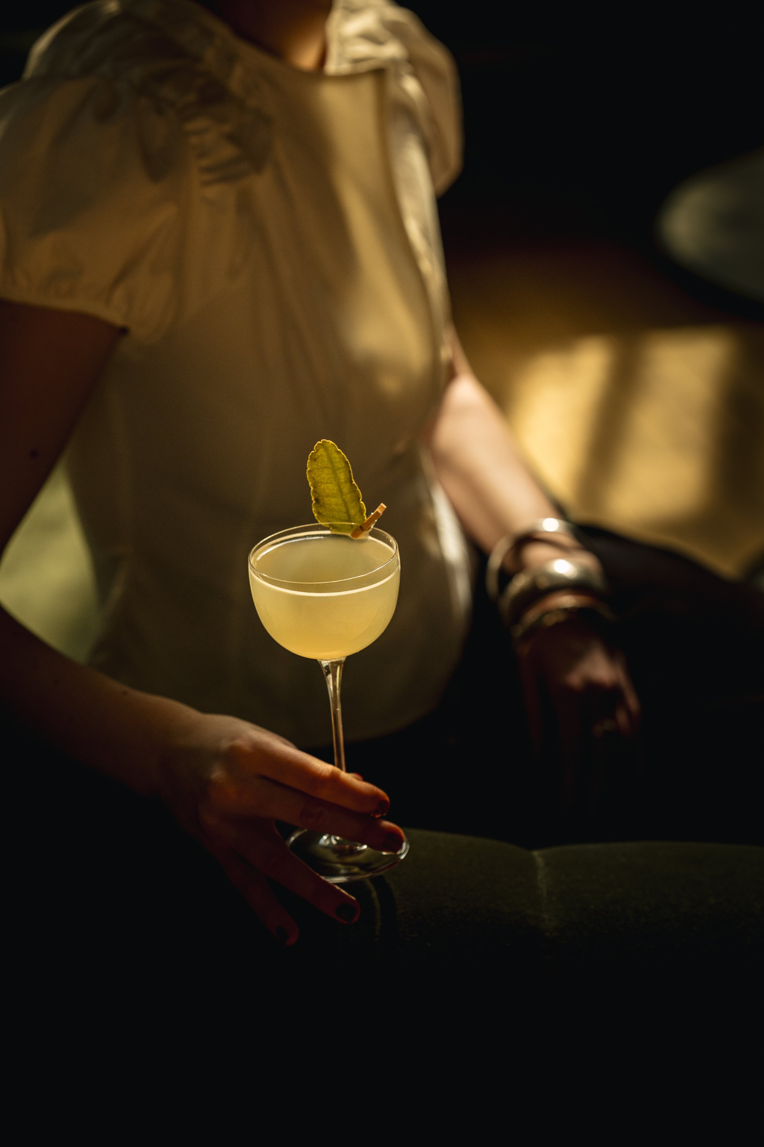 A person holding a cocktail glass with a yellow drink and a small yellow leaf garnish. The person is wearing a yellow top and jewelry, and the background is dark.