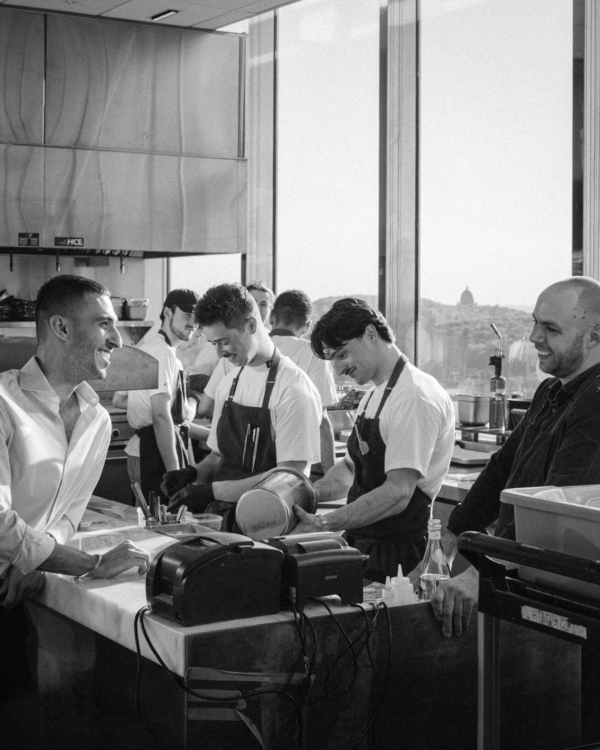 A group of young men and older man standing around a table in a well-lit kitchen or cafeteria, engaging with each other and food. In the background, a large window shows a distant building or monument on a hill.