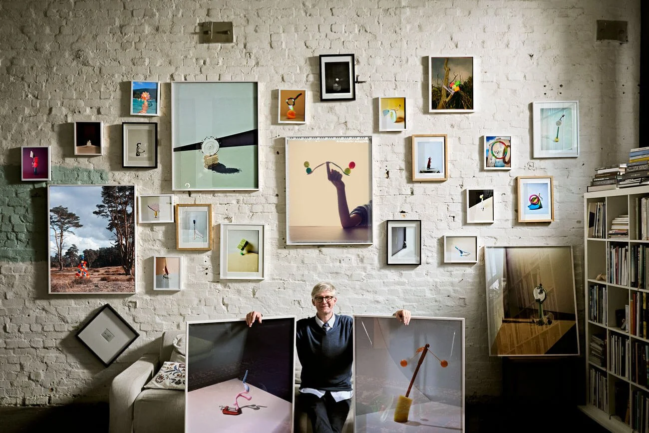 Der Gründer von Highlevelzero sitzt in einem Raum vor einer Wand, die mit vielen Bilderrahmen von verschieden großen Fotos und Bildern dekoriert ist. Es gibt auch ein Regal mit Büchern.