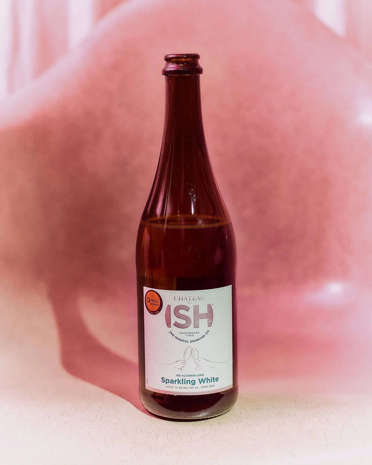 Eine Flasche alkoholfreier Sparkling White Wein der Marke Château ISH, fotografiert vor einem verschwommenen rosa Hintergrund.