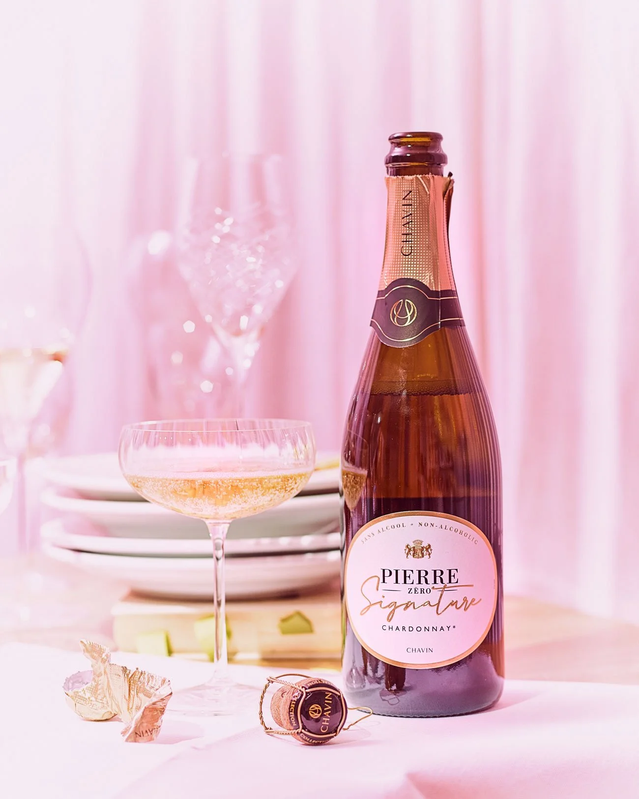 Flasche alkoholfreier Chardonnay Pierre Zero Signature mit Champagnerglas auf einem Tisch, dekoriert mit gefüllten Schokoladenpapieren und gestapelten Tellern im Hintergrund.