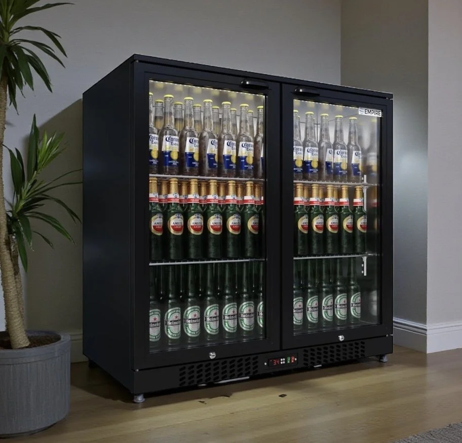 A black beverage refrigerator filled with bottles of beer, including Corona, Amstel, and Heineken, placed in a room with a potted plant nearby.