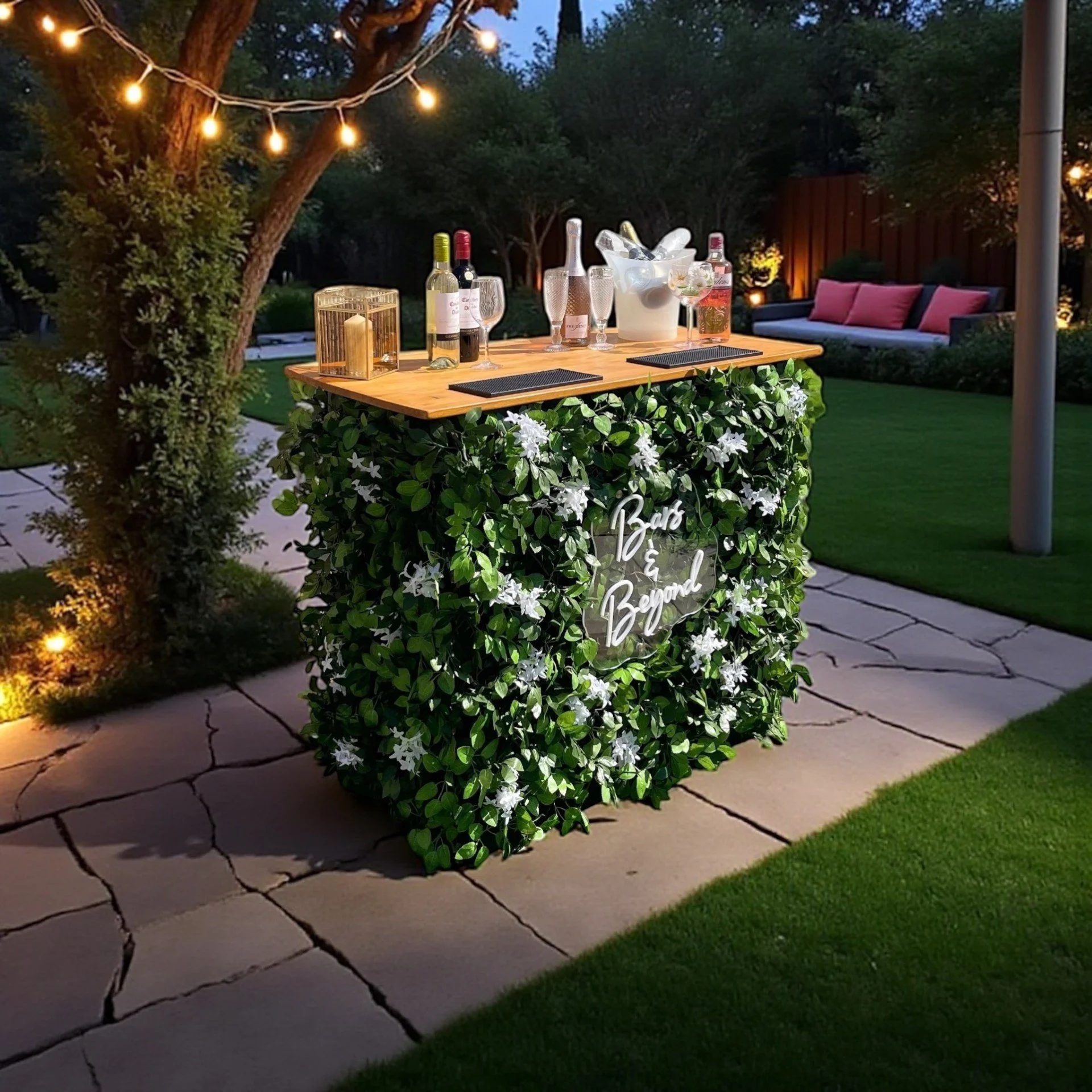 A backyard patio with a decorated bar area for outdoor gathering, featuring a flowery green bush with a white sign that reads 'Bar & Beyond', a wooden table with wine bottles, glasses, and an ice bucket, a seating area with cushions, a wooden fence, trees, and string lights illuminating the scene at dusk.