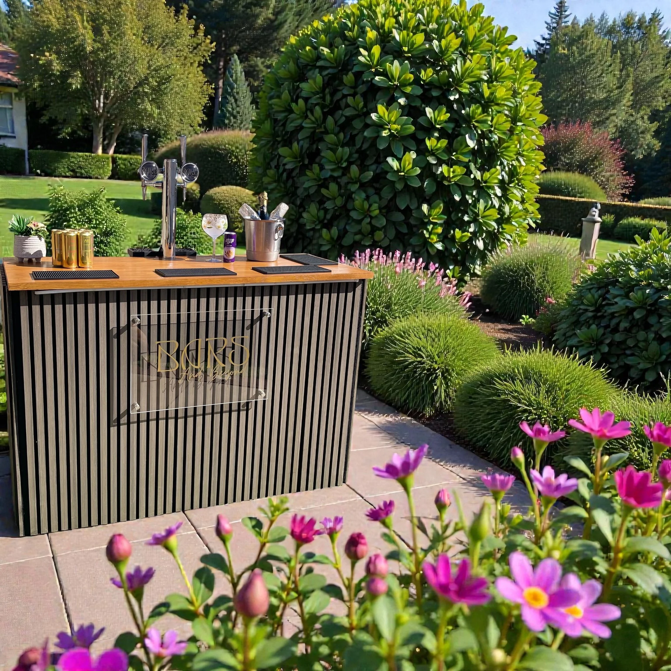 Outdoor bar in a garden setting with lush green trees and bushes, a large leafy shrub, pink and purple flowers, and a wooden bar with drink cans, a glass of drink, and bartending tools on top.