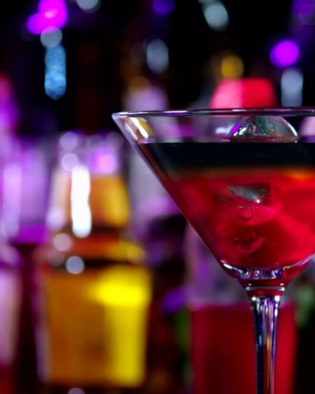 A close-up of a red cocktail with a lime wedge in a martini glass, with colorful blurred bottles and bokeh light effects in the background.