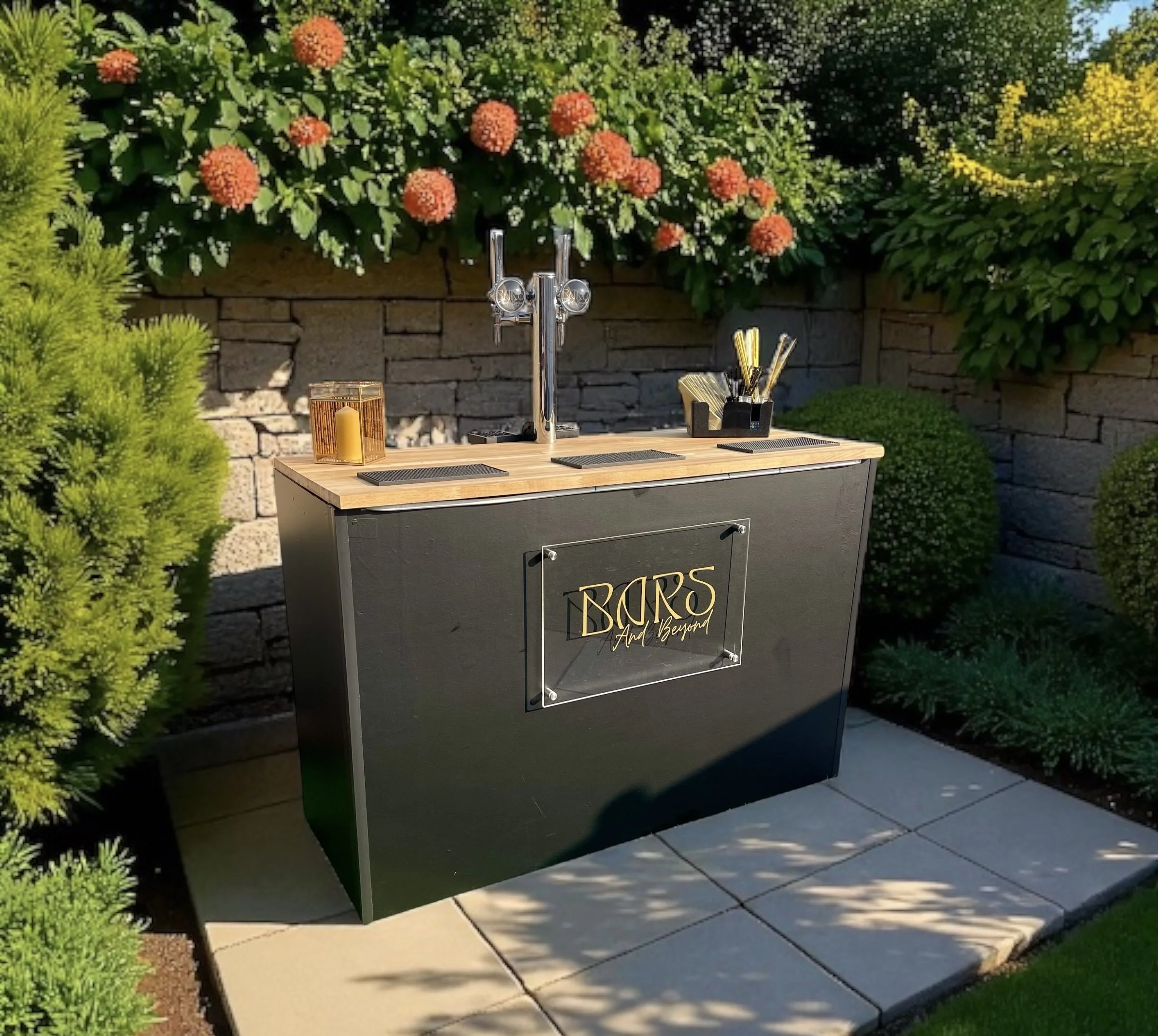 Outdoor bar setup with a wooden countertop next to a stone wall, surrounded by greenery and colorful flowering plants.