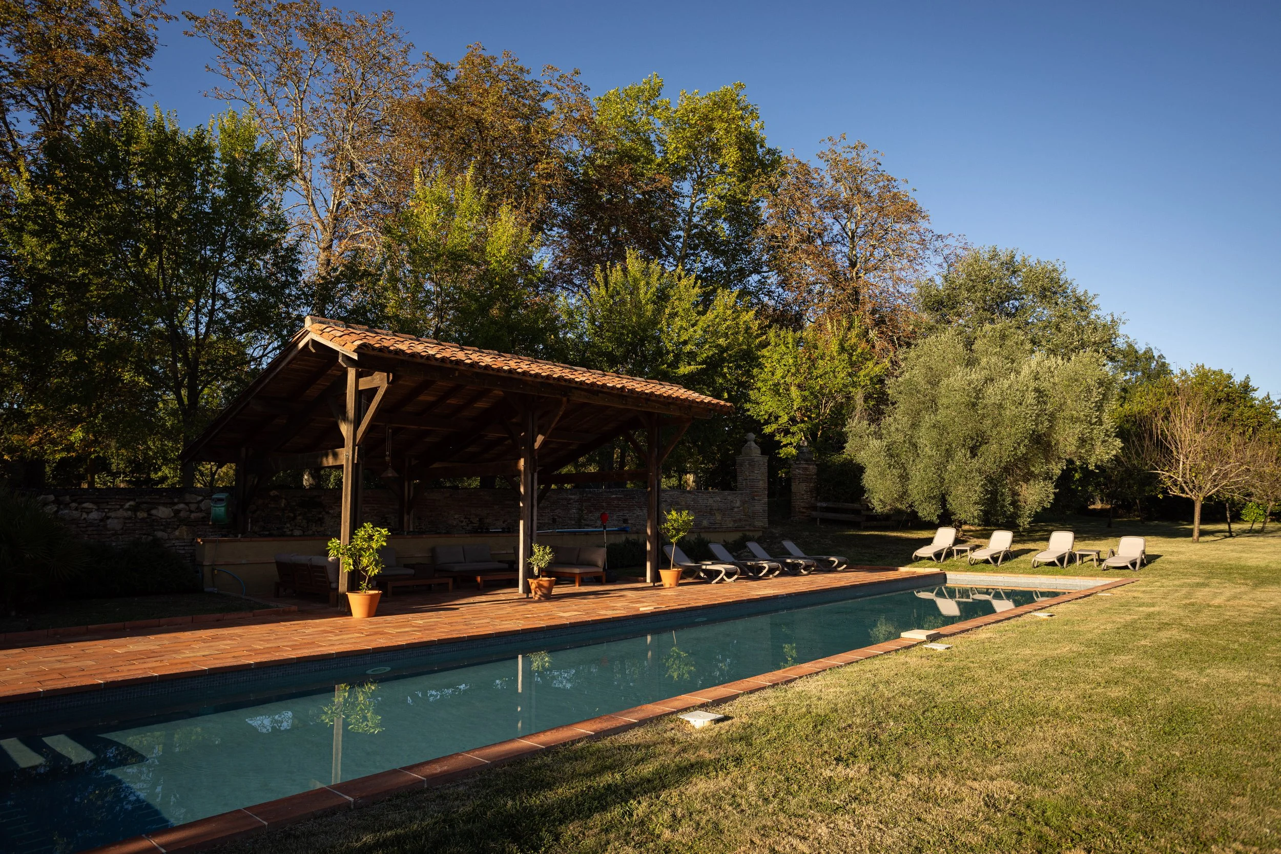 Outdoor swimming pool at Château de Sirengard, surrounded by landscaped gardens, mature trees, sun loungers, and a shaded poolside pavilion in the French countryside.