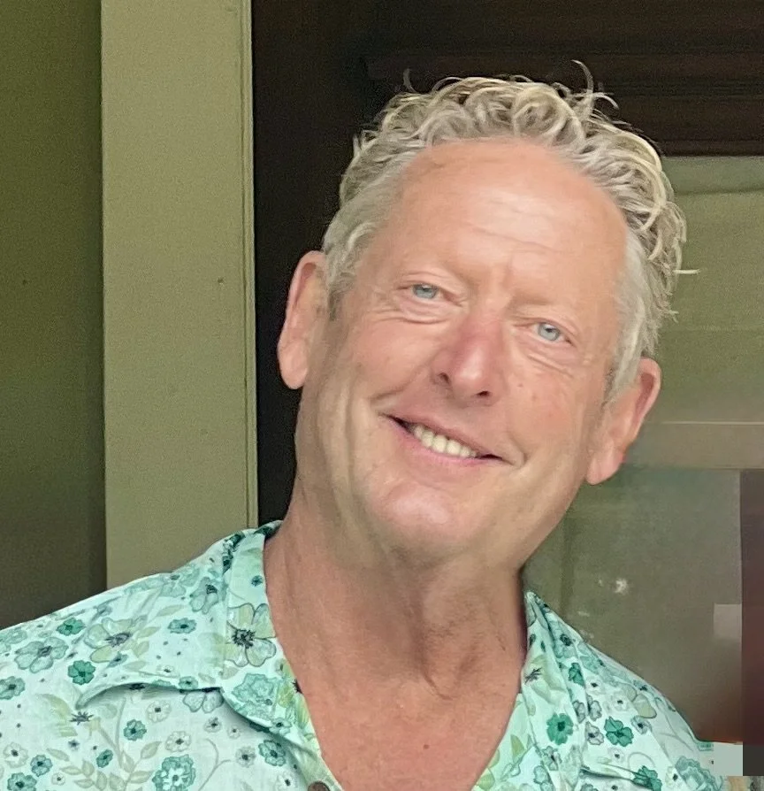 A smiling man with curly gray hair, wearing a light green shirt with a floral pattern, standing in front of a dark door and green wall.
