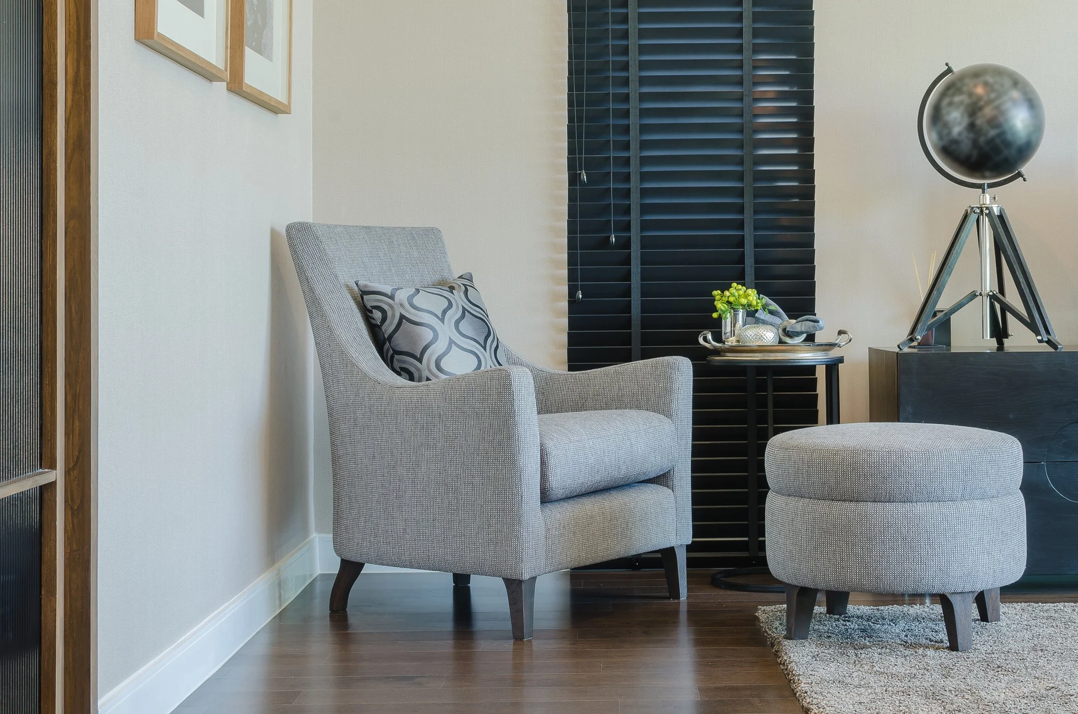 A cozy living room corner with a light gray armchair, a round matching ottoman, a small black side table with a vase of green flowers, and a black globe on a tripod stand, near a window with black blinds.