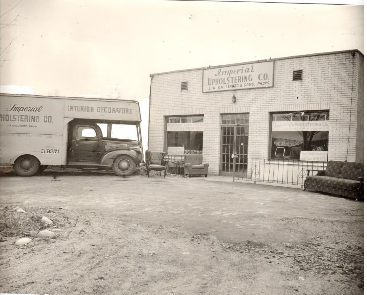 Black and white photograph of an old storefront named Imperial Upholstering Co, with signage indicating furniture and upholstery services. In front, there are vintage chairs and sofas, and an old truck with writing on its side parked beside the building.