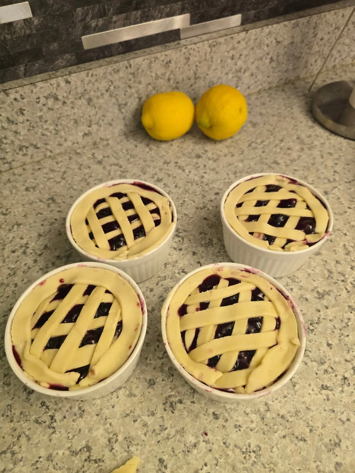 Four small ramekins with uncooked lattice-top pies on a kitchen countertop, with two lemons in the background.