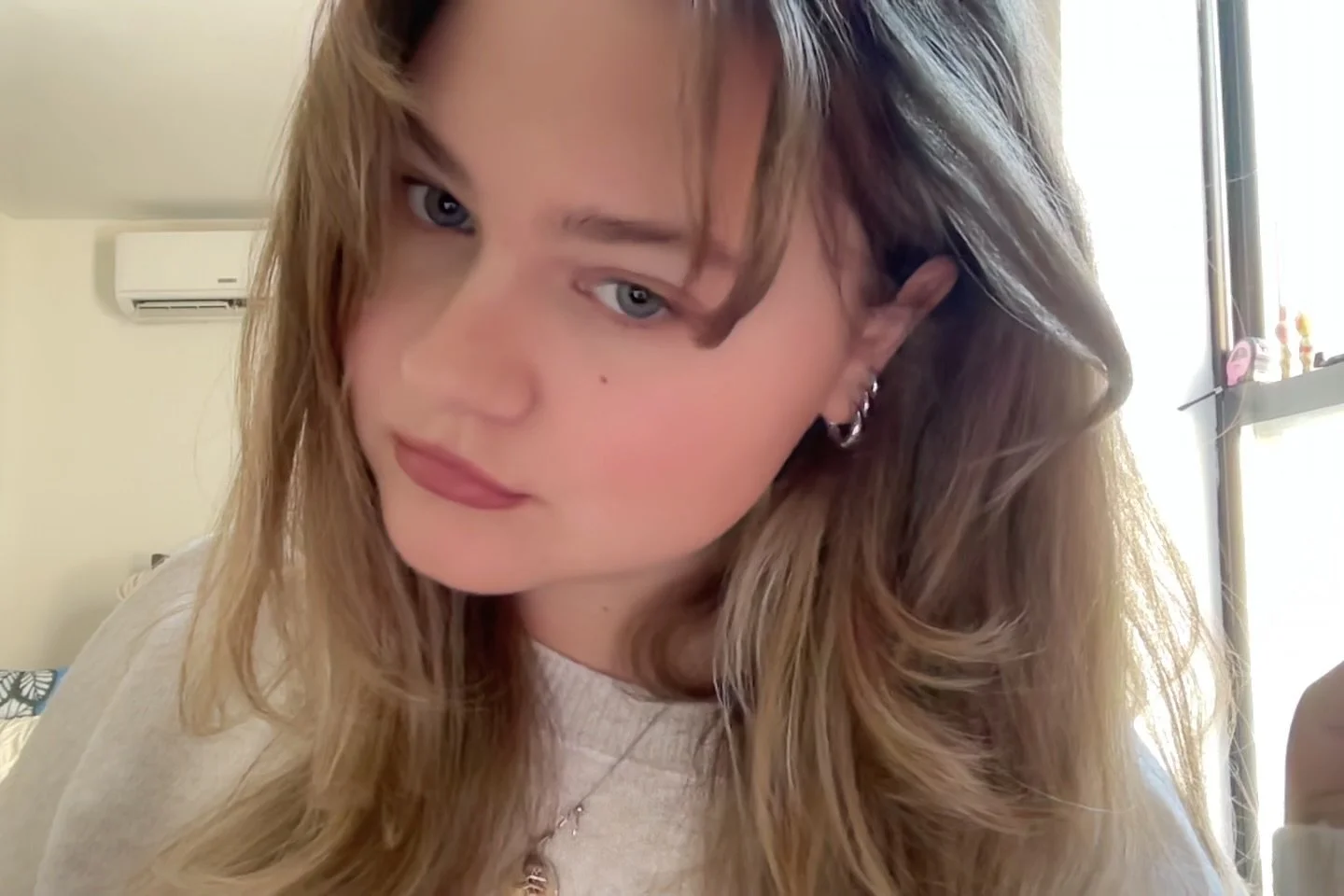 A woman with long wavy hair and earrings looks into the camera indoors near a window, with an air conditioner on the wall behind her.