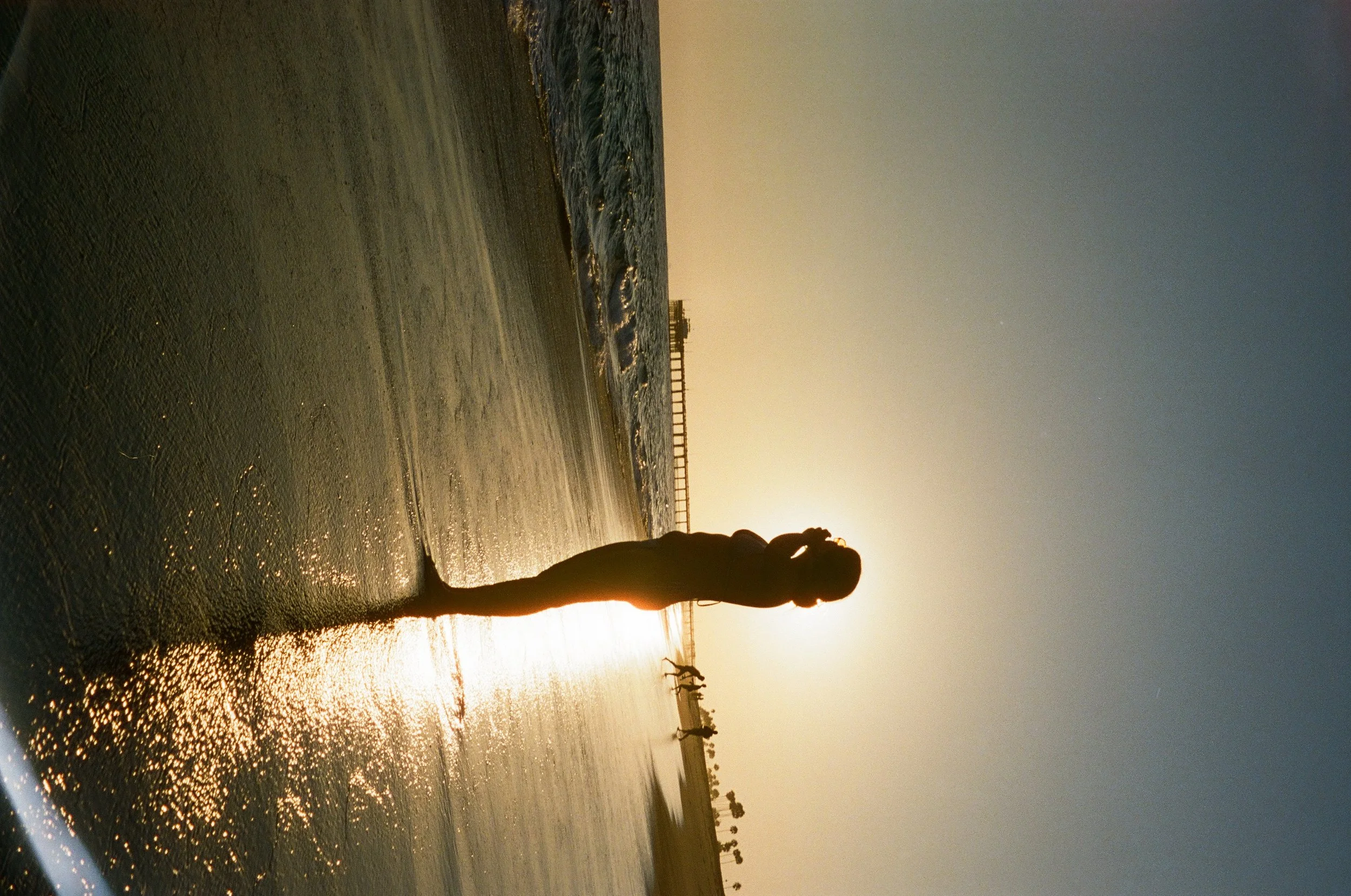 A person standing in the shallow water of a beach at sunset, with the sun low on the horizon and a wooden pier in the distance.