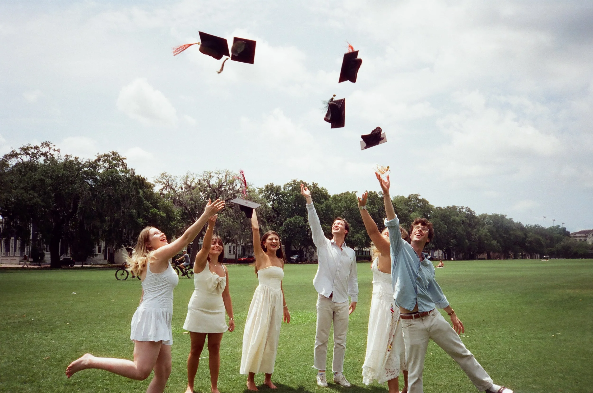 Group of young adults celebrating graduation outdoors, throwing caps in the air on a grassy field with trees and cloudy sky in the background.