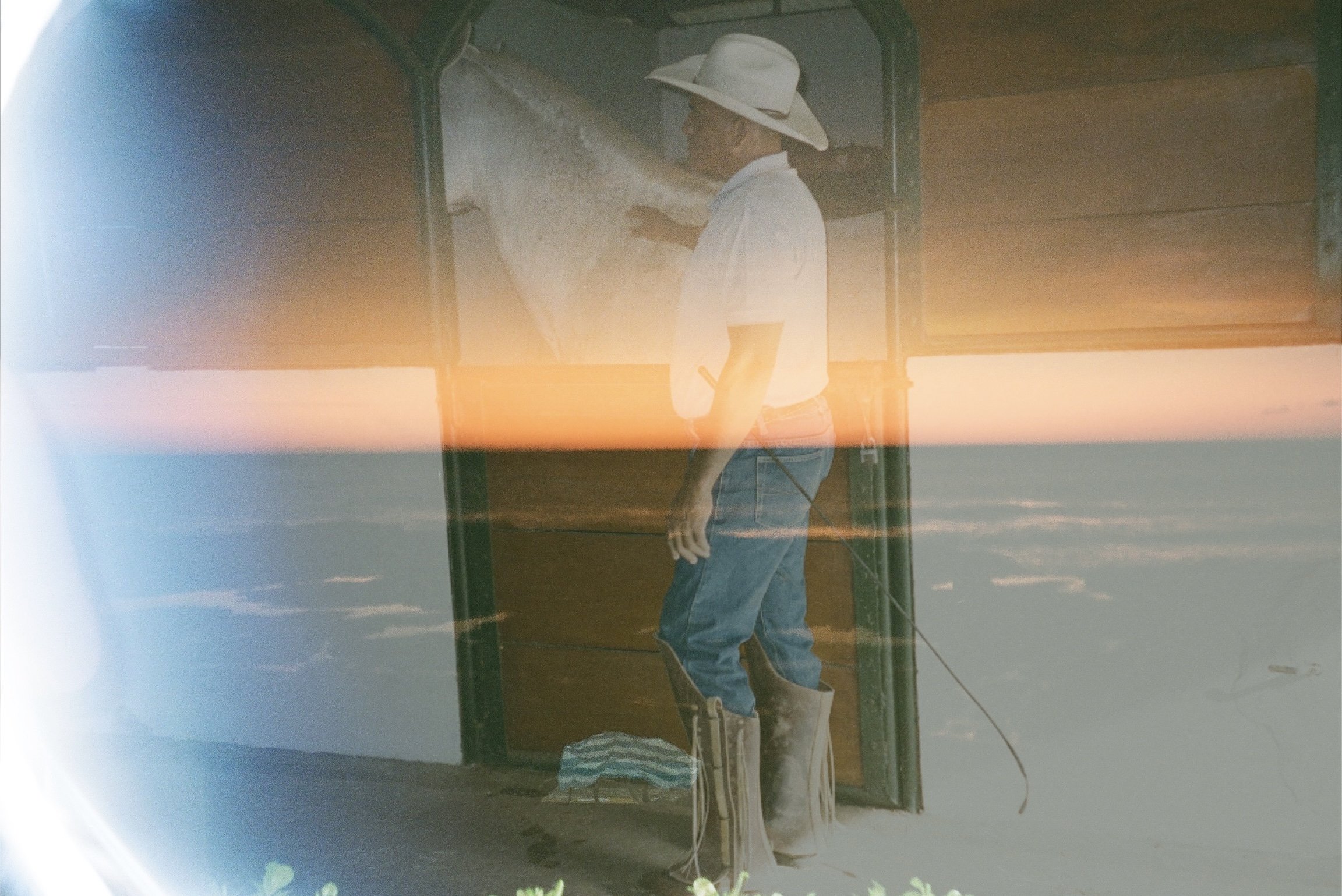 A woman wearing a wide-brimmed hat, white shirt, and jeans appears to be fishing, standing on a boat at sunset with calm water around her.