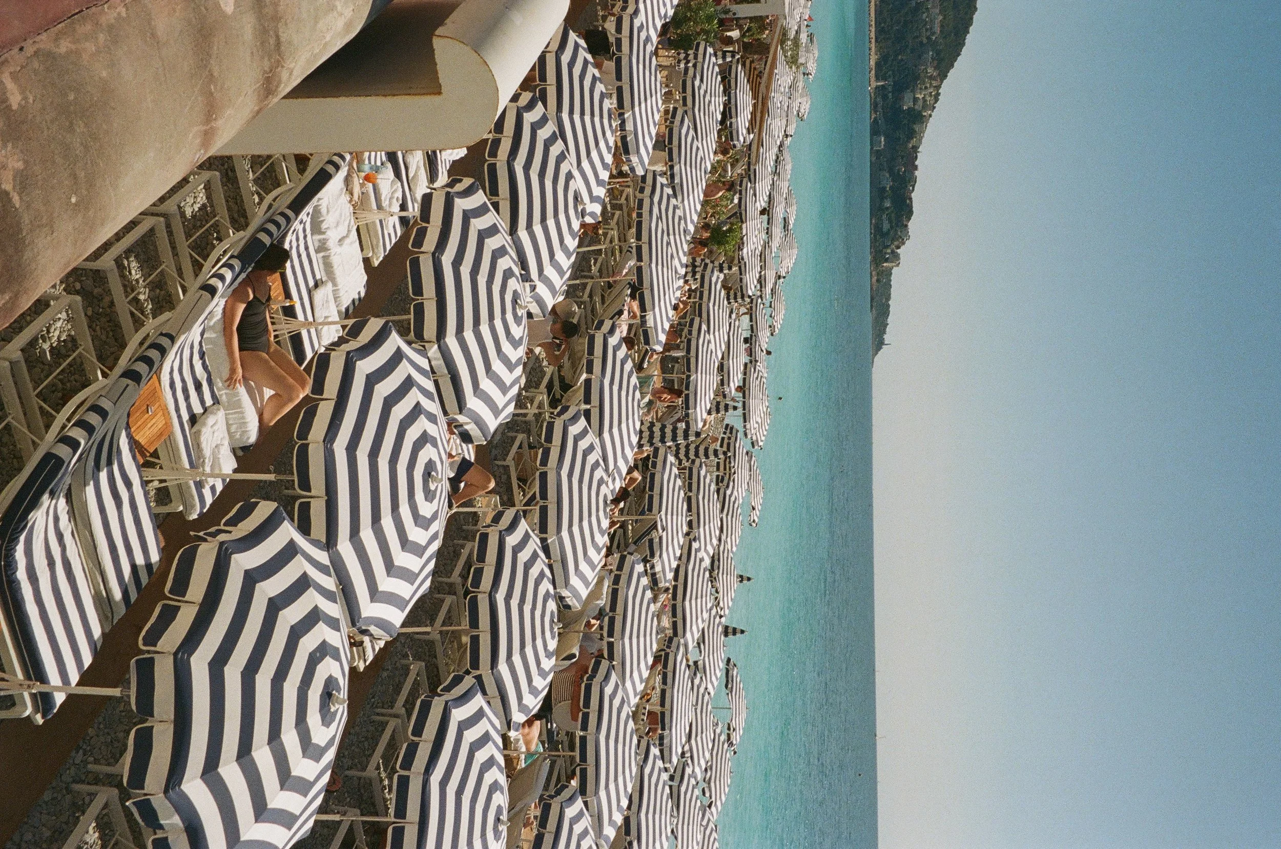 Beach with numerous striped blue and white lounge chairs and umbrellas near the shoreline, with people relaxing and enjoying the sun and ocean in the background.